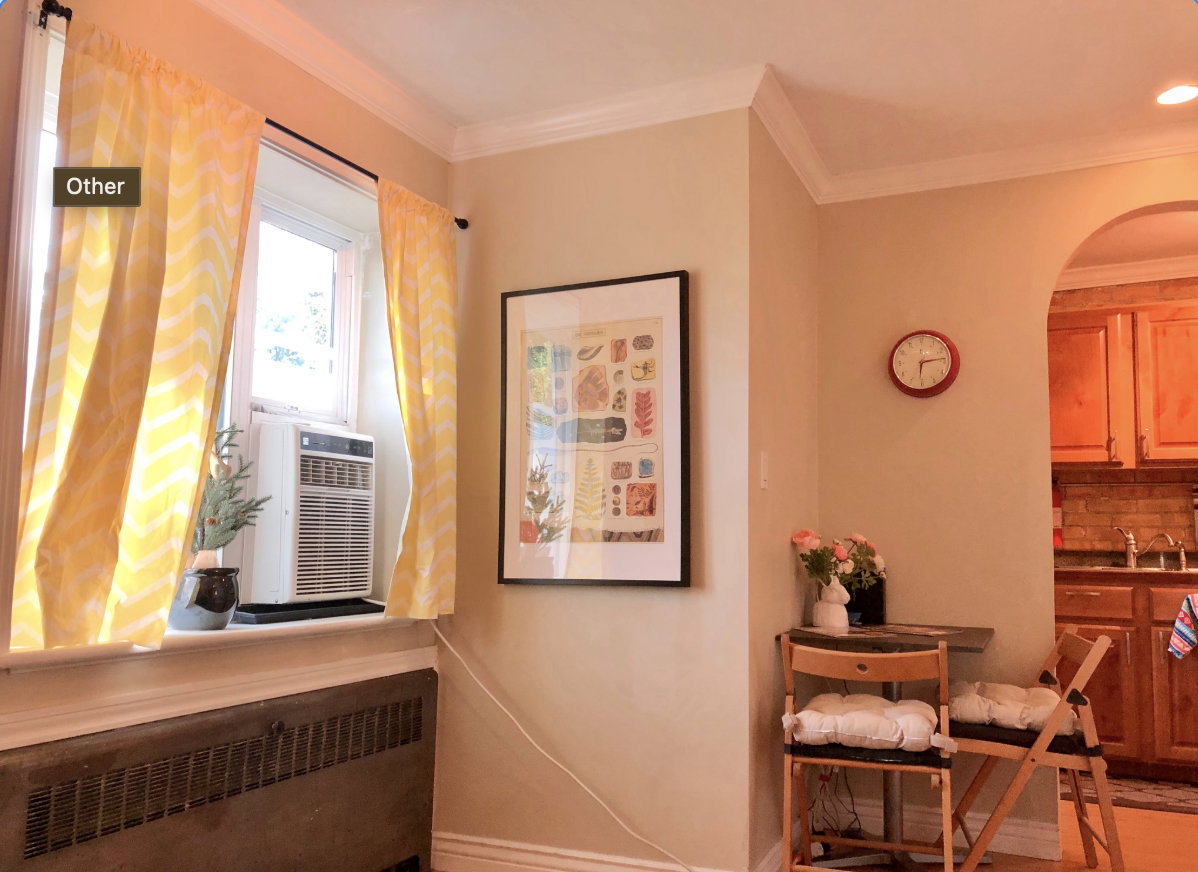 Crown molding and neutral wall paint define this living space, featuring a window with natural light, a baseboard radiator, and an arched doorway leading into a kitchen with wood cabinetry and a tile backsplash