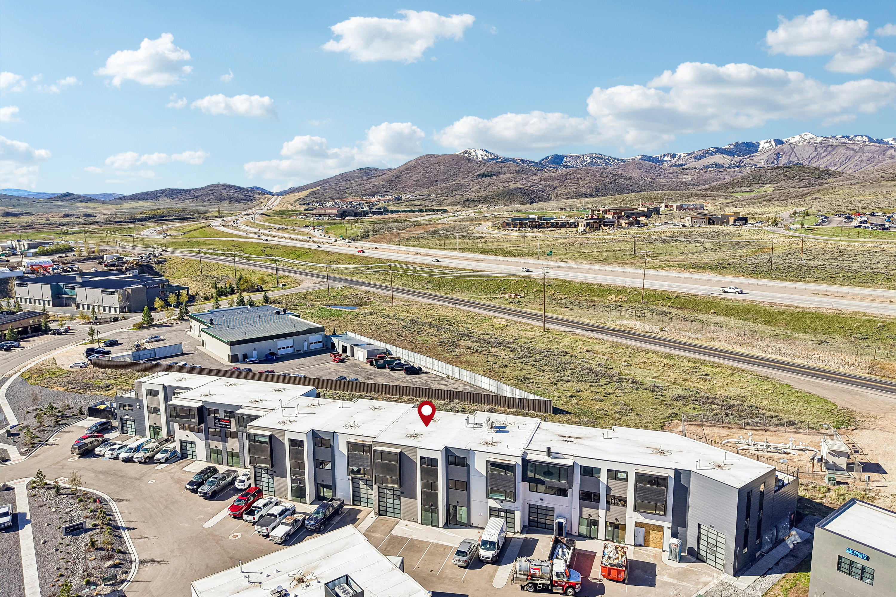 Modern commercial building with a grey and white facade, multiple roll-up garage doors, ample on-site parking, and immediate proximity to a major highway