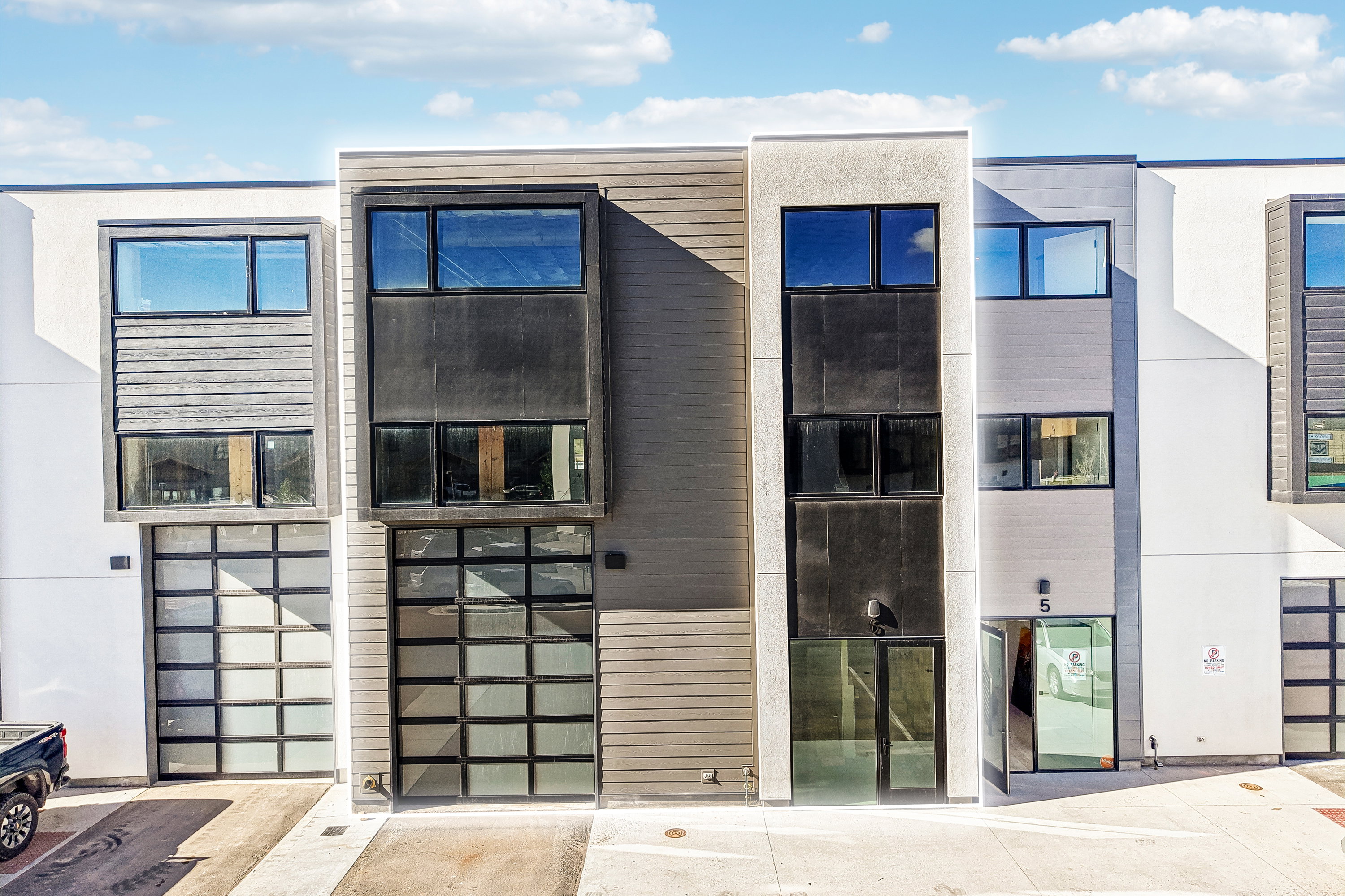 Modern townhome facade featuring a prominent glass-paneled garage door, large rectangular windows, and a multi-textured exterior combining light-colored stucco and dark horizontal siding