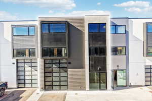 Modern townhome facade featuring a prominent glass-paneled garage door, large rectangular windows, and a multi-textured exterior combining light-colored stucco and dark horizontal siding