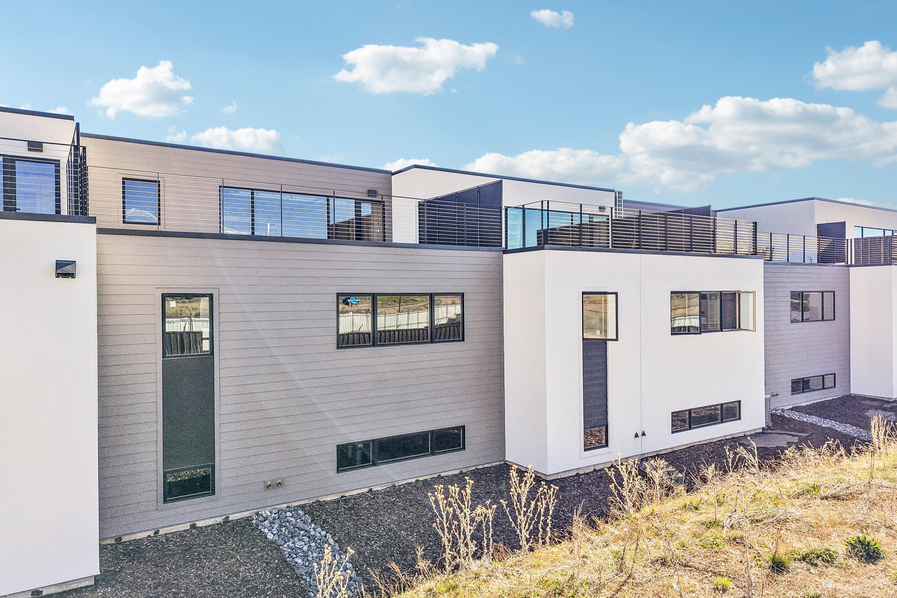 Modern multi-level exterior featuring white and grey siding, black-framed windows, and rooftop decks with glass and slatted railings