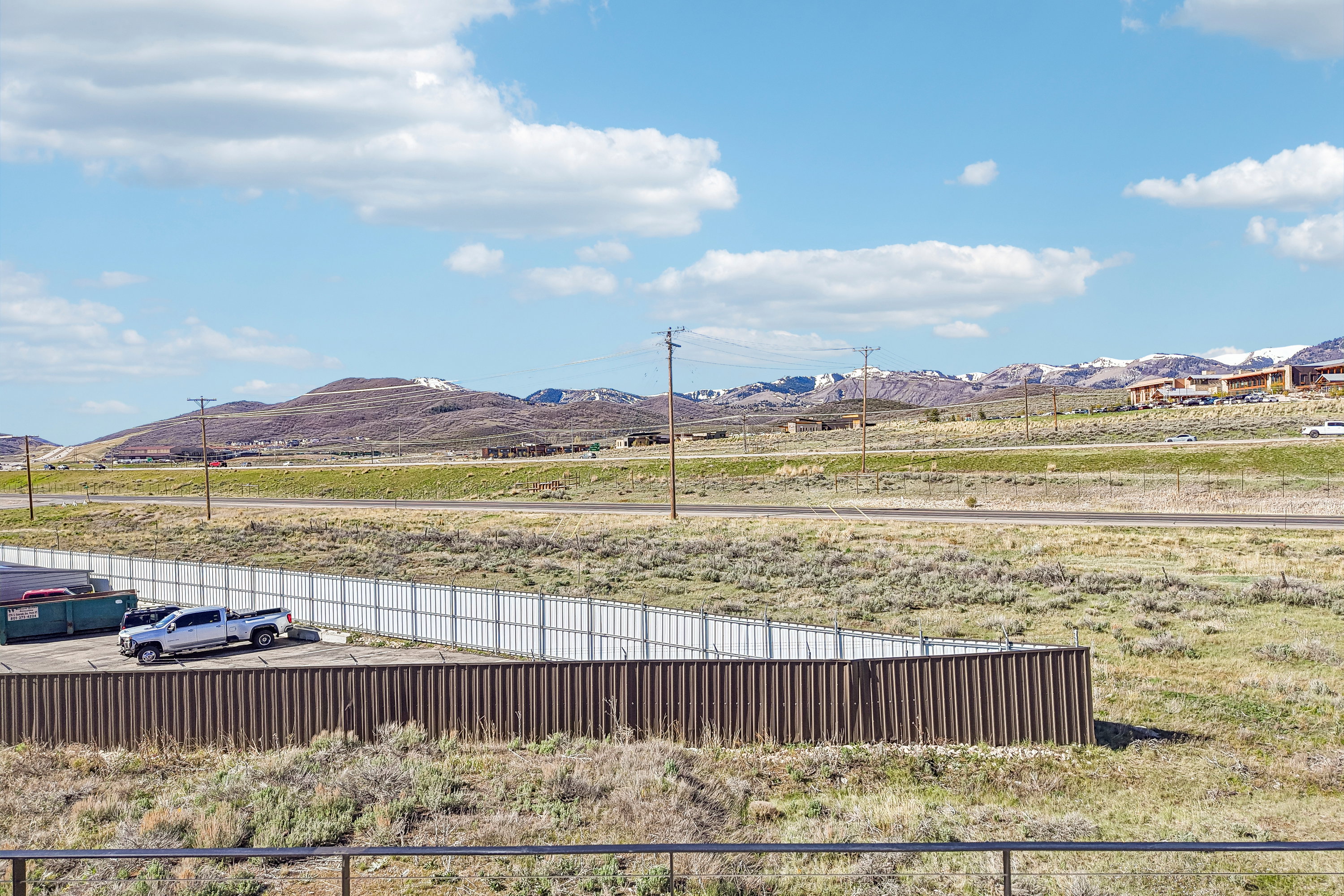 Expansive undeveloped land featuring a partial perimeter fence, a brown corrugated metal retaining wall, and distant snow-capped mountains
