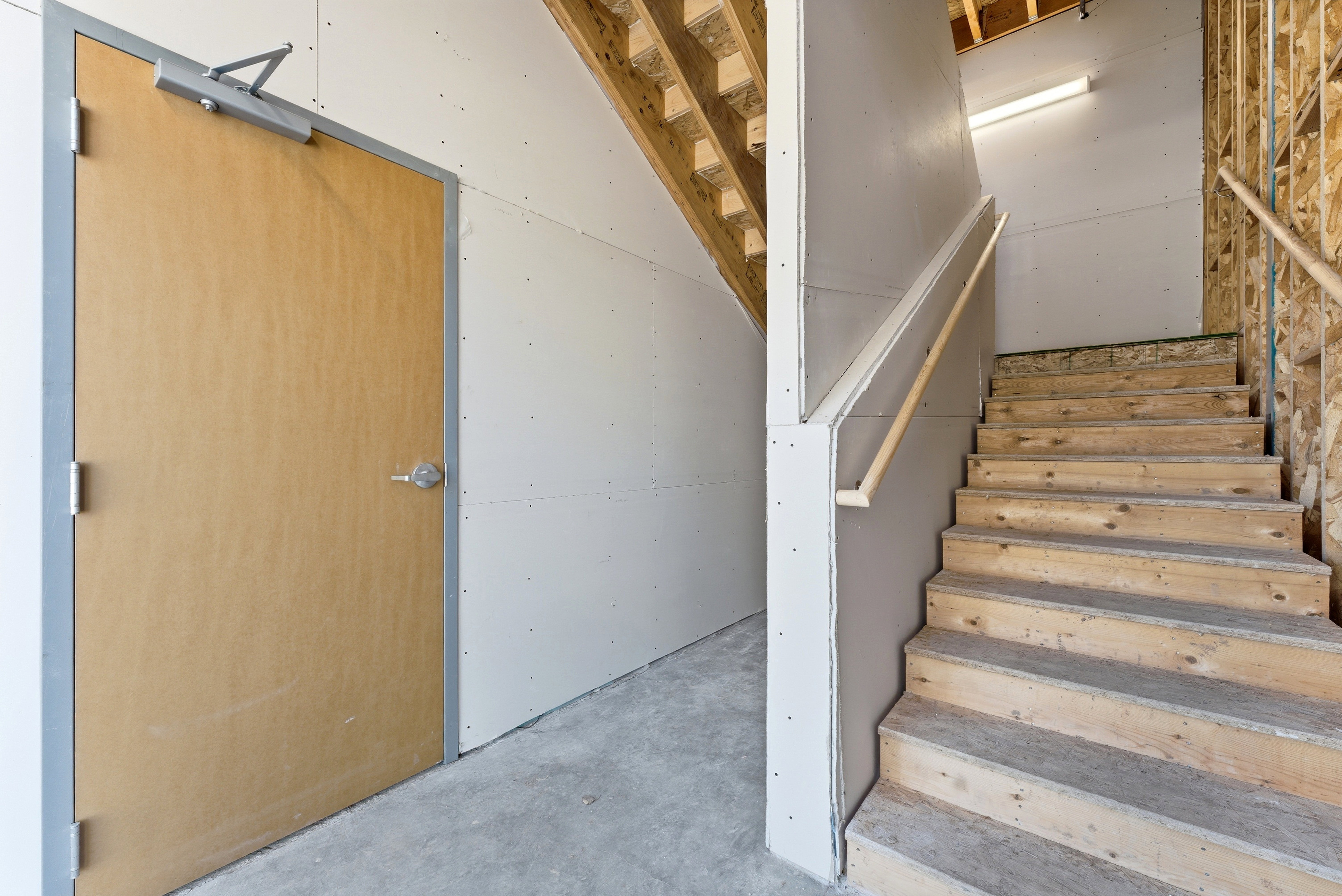 Unfinished interior space featuring a light wood-finish door with metal frame, wood staircase with handrail, exposed wood framing, unpainted drywall, and a concrete floor