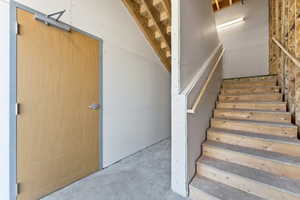 Unfinished interior space featuring a light wood-finish door with metal frame, wood staircase with handrail, exposed wood framing, unpainted drywall, and a concrete floor