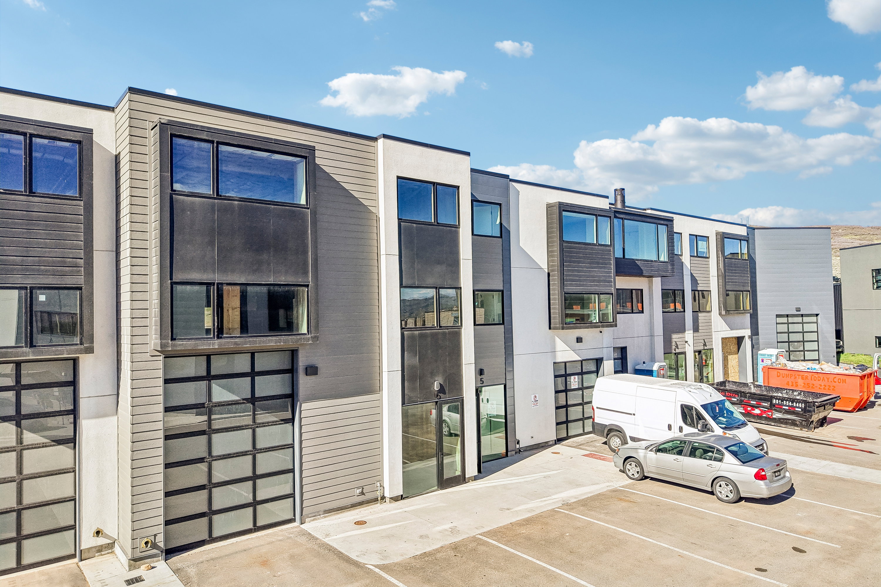 Modern multi-level exterior featuring light gray horizontal siding, dark accent panels, large windows, and contemporary glass panel garage doors