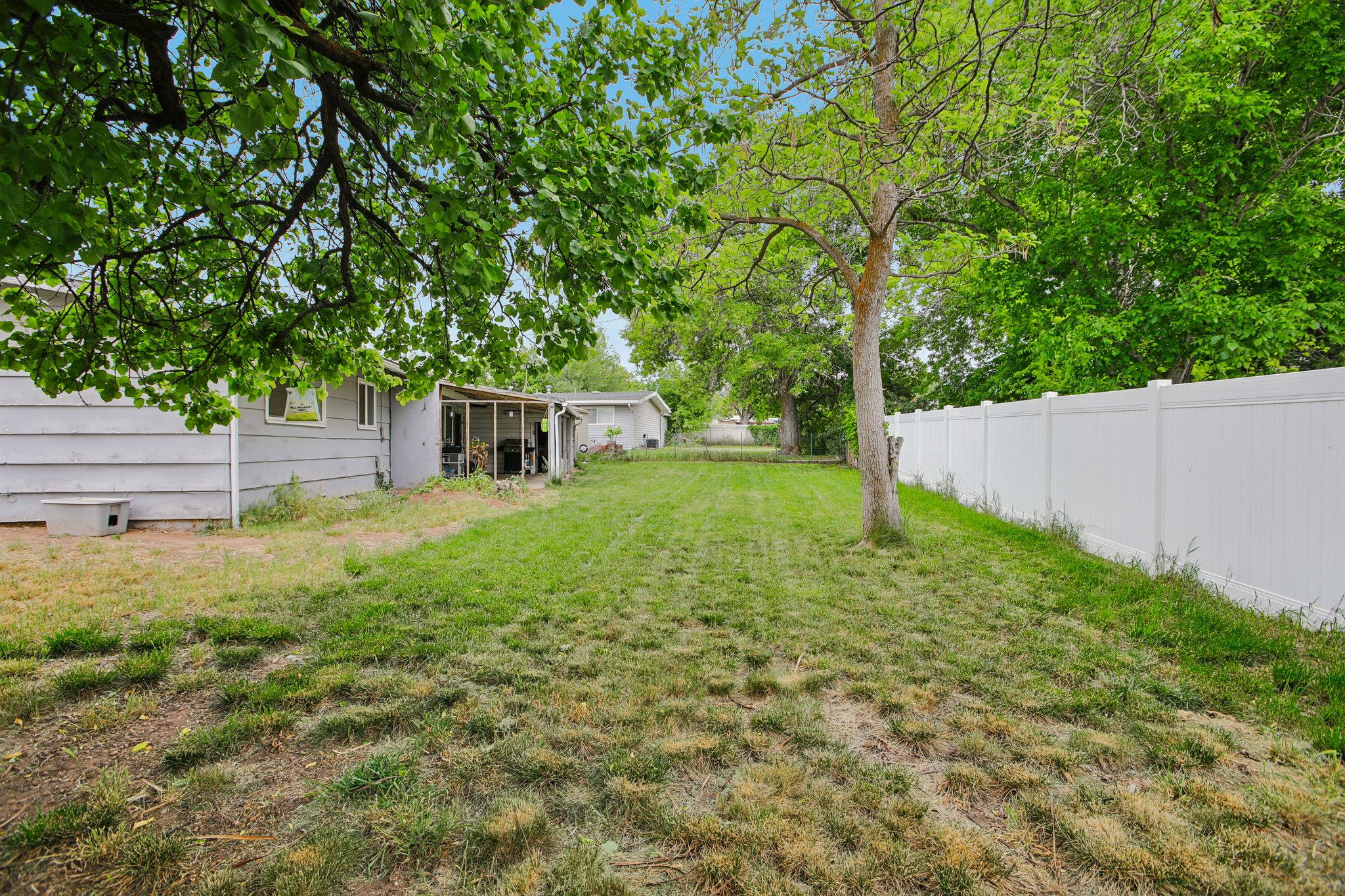 Expansive backyard featuring a white privacy fence, mature trees, and a covered patio area