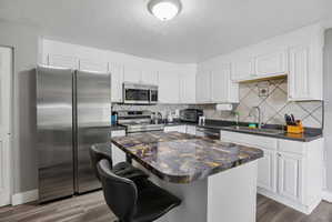 Kitchen featuring a central island with a multi-toned countertop, white cabinetry, stainless steel appliances, a tiled backsplash, and wood-finish flooring