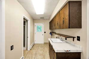 Utility space featuring tile flooring, a white porcelain sink, tile countertops, wood-finish cabinetry, and a full-light exterior door