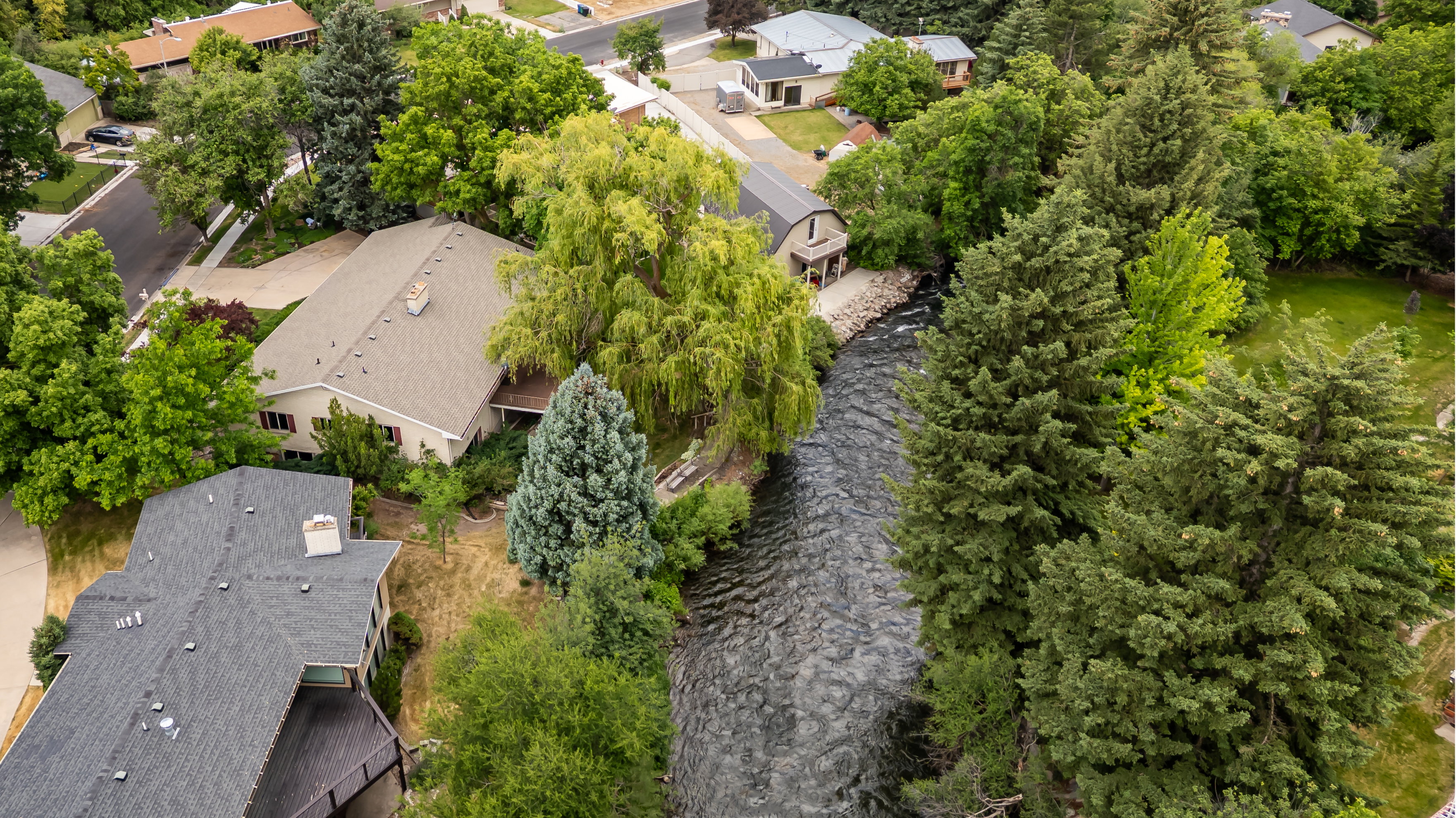 Riverfront property featuring a stone and siding exterior, mature trees, a private deck, and a shingle roof