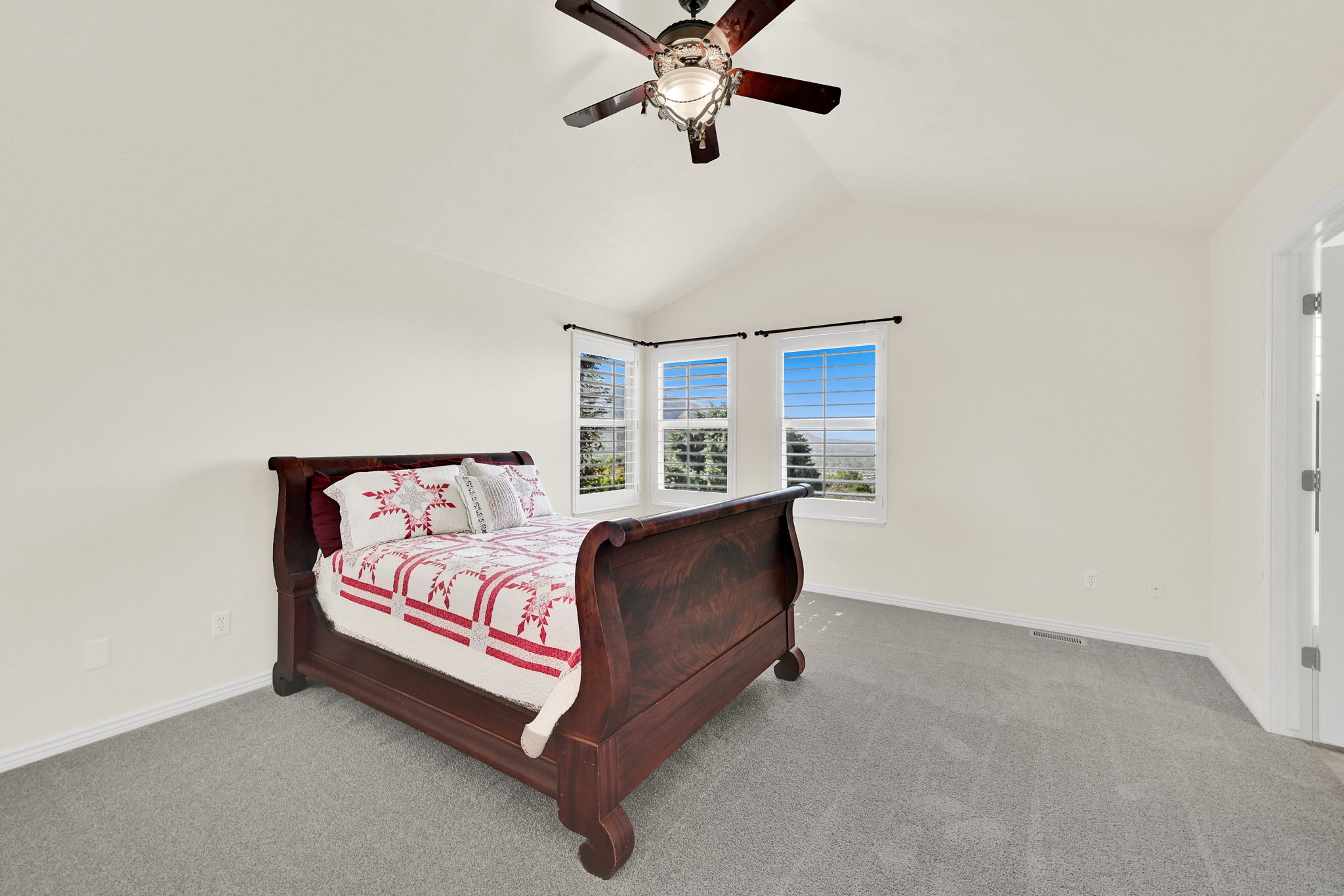 Vaulted ceiling with a ceiling fan, three windows with plantation shutters, light-colored walls, and gray carpet flooring