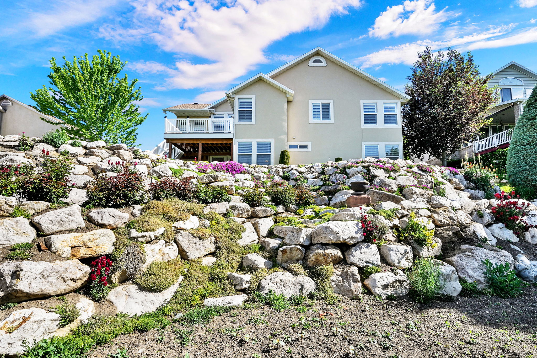 Rear exterior featuring a multi-level rock retaining wall with diverse plantings, a second-story balcony with white railing, and a stucco finish