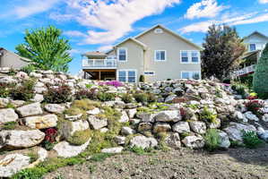 Rear exterior featuring a multi-level rock retaining wall with diverse plantings, a second-story balcony with white railing, and a stucco finish