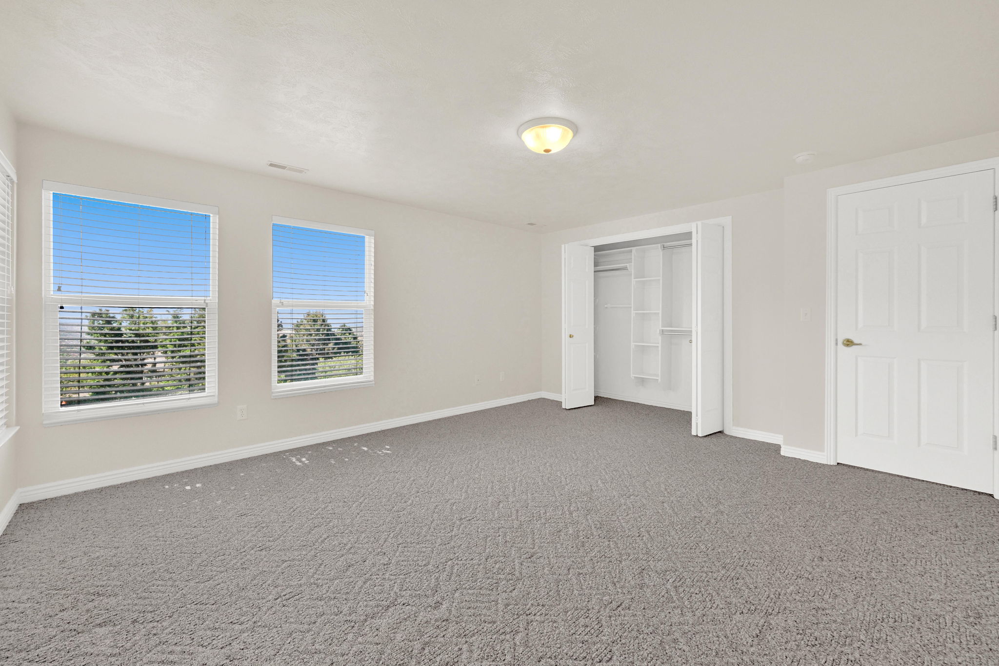 Spacious room with two large windows featuring horizontal blinds, neutral wall paint, gray patterned carpet, a built-in closet with shelving, and a six-panel door