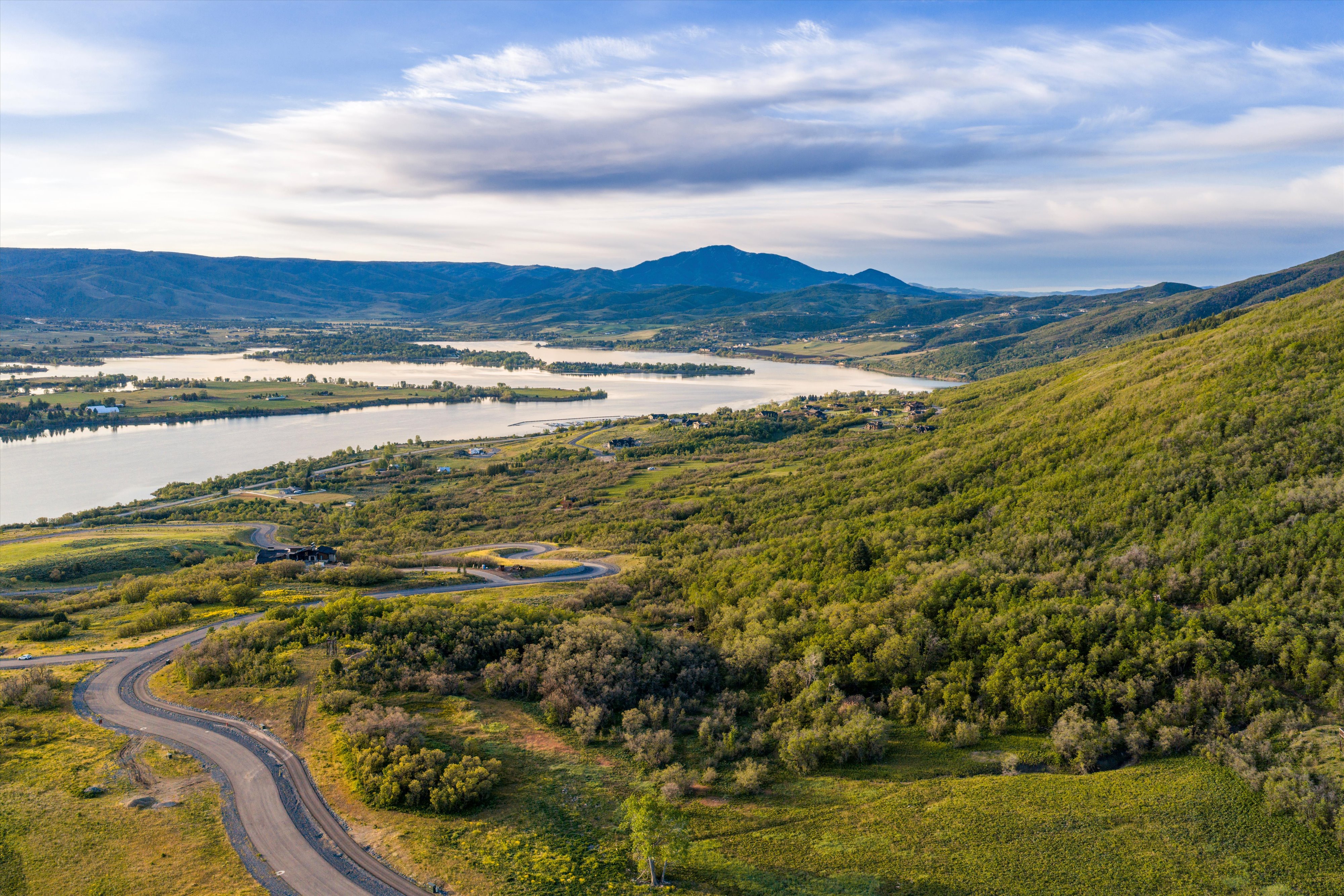 Expansive natural landscape featuring a winding paved road, lush green hillsides, a large body of water, and distant mountains