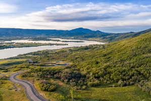 Expansive natural landscape featuring a winding paved road, lush green hillsides, a large body of water, and distant mountains