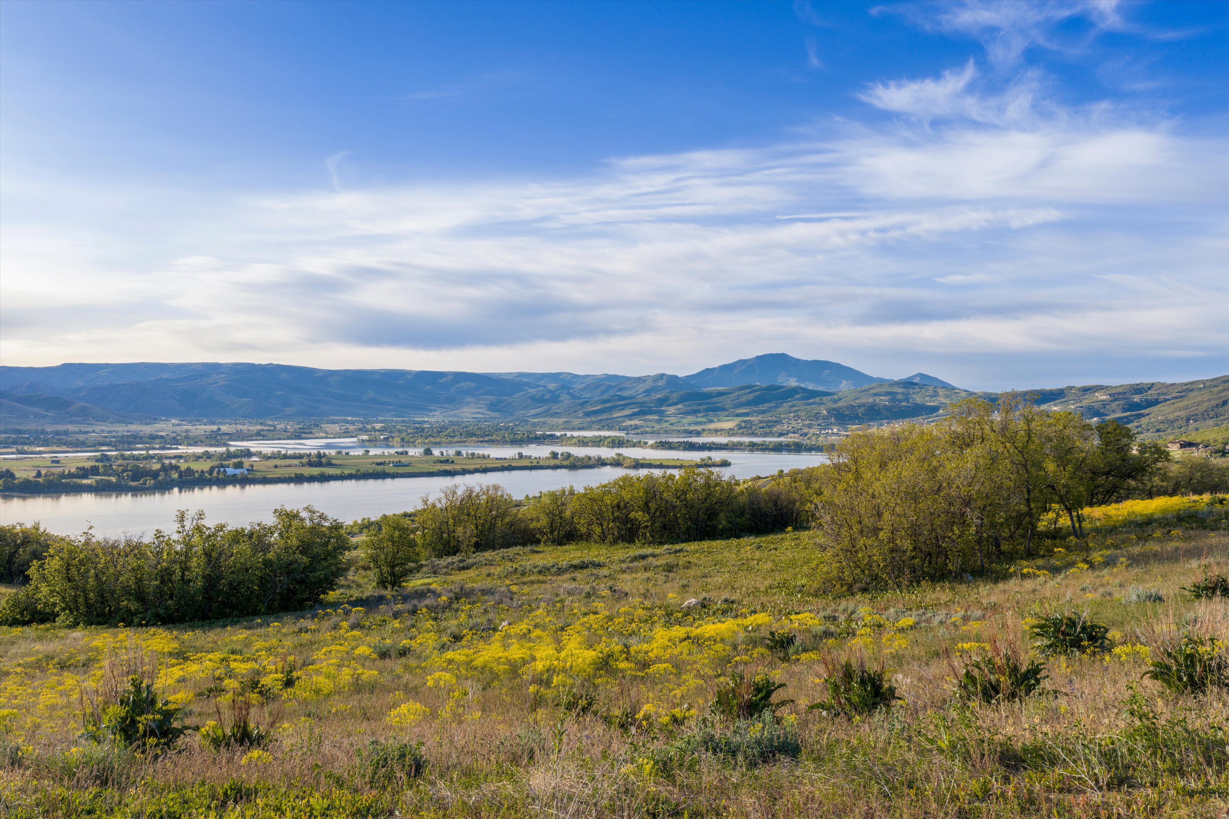 Expansive natural landscape featuring rolling hills with green and yellow foliage, a prominent body of water, distant mountains, and a clear blue sky
