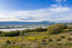Expansive natural landscape featuring rolling hills with green and yellow foliage, a prominent body of water, distant mountains, and a clear blue sky