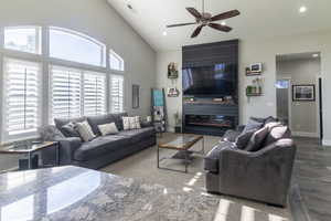 Vaulted ceiling living area featuring a prominent fireplace surround with built-in shelving, wood-finish flooring, a ceiling fan, and large windows with integrated plantation shutters
