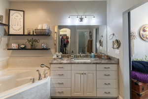 Bathroom featuring a built-in corner bathtub with deck-mounted faucet, a vanity with under-mount sink and brushed nickel faucet, a large wall-mounted mirror, a three-light vanity fixture, and open shelving