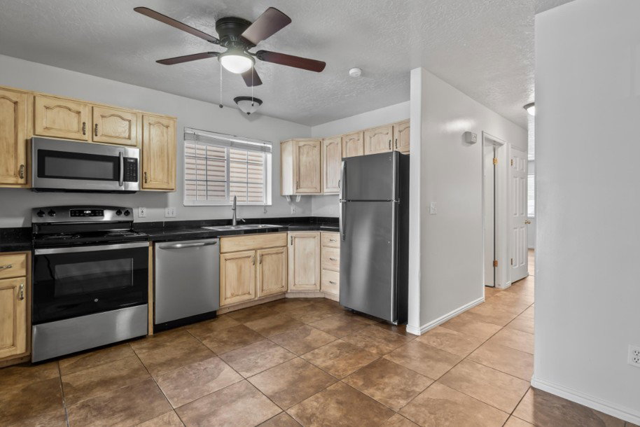 Kitchen featuring wood-finish cabinetry, stainless steel appliances, black countertops, and a ceiling fan with integrated lighting
