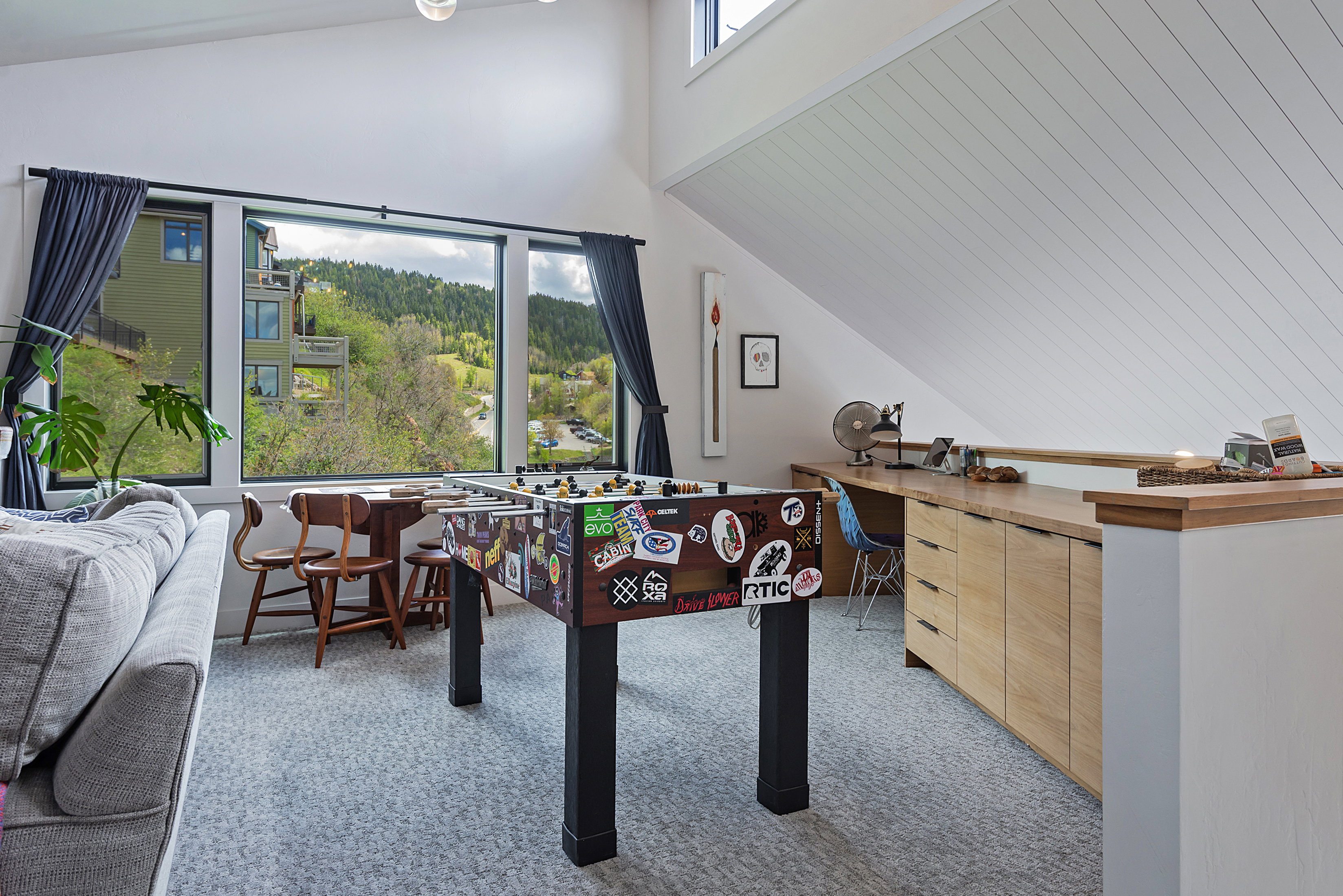 Expansive window wall with black trim, built-in desk with wood-finish cabinetry, vaulted ceiling with wood paneling, and light gray patterned carpeting