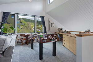 Expansive window wall with black trim, built-in desk with wood-finish cabinetry, vaulted ceiling with wood paneling, and light gray patterned carpeting