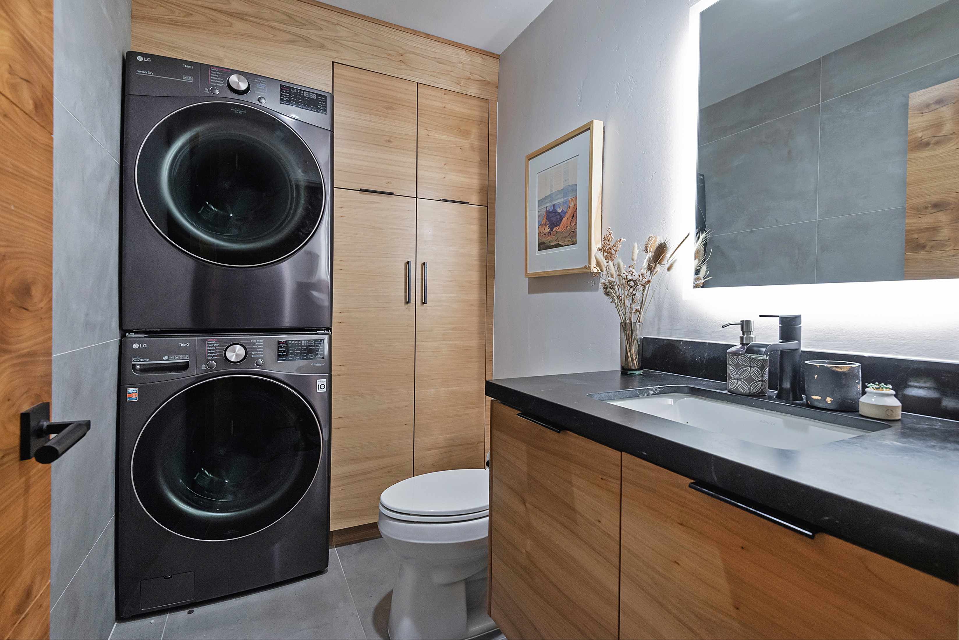 Compact laundry area with stacked front-load machines, a single-sink vanity with a dark countertop, wood-finish cabinetry, and an illuminated mirror