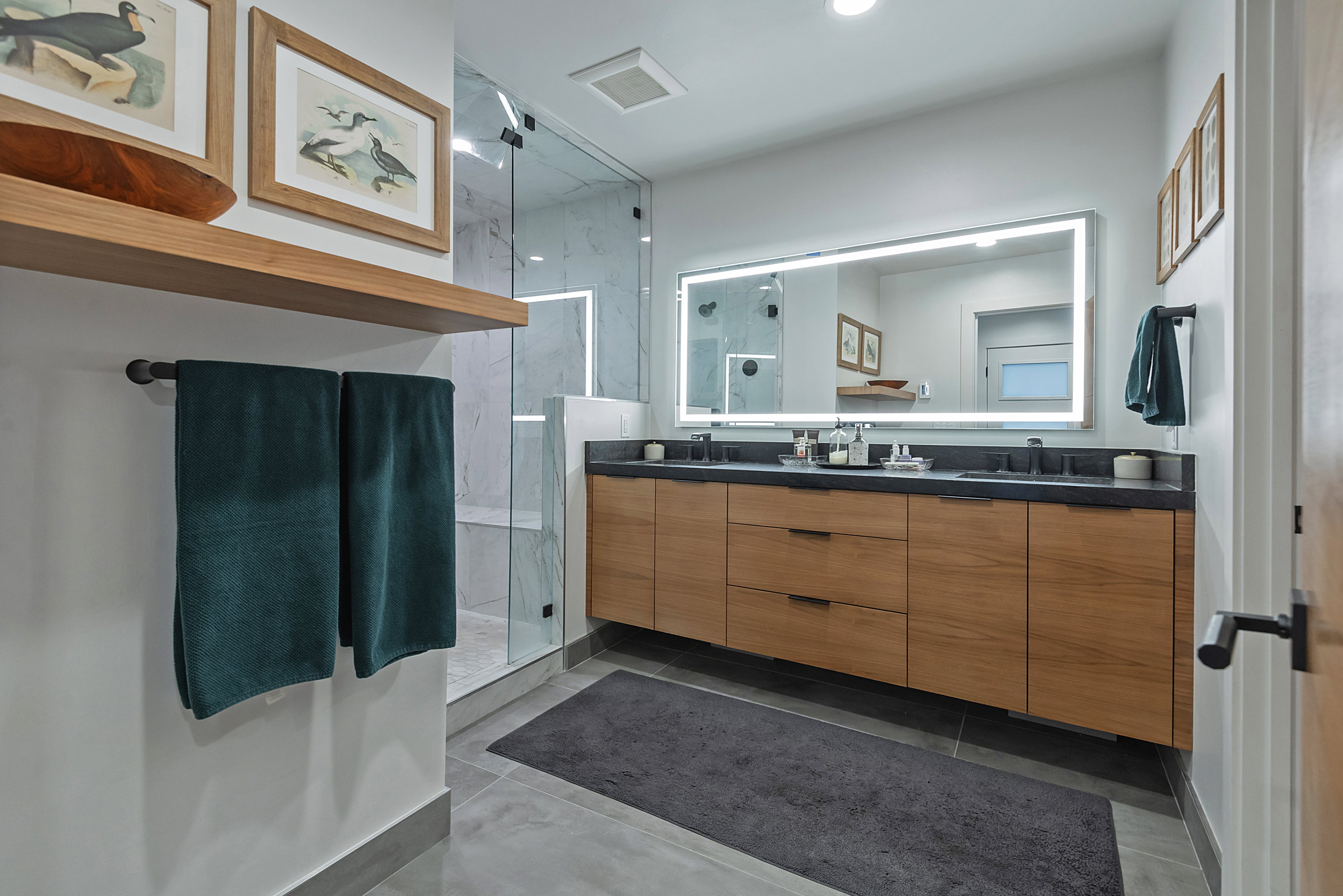 Contemporary bathroom featuring a dual vanity with wood-finish cabinetry, a large illuminated mirror, and dark countertops