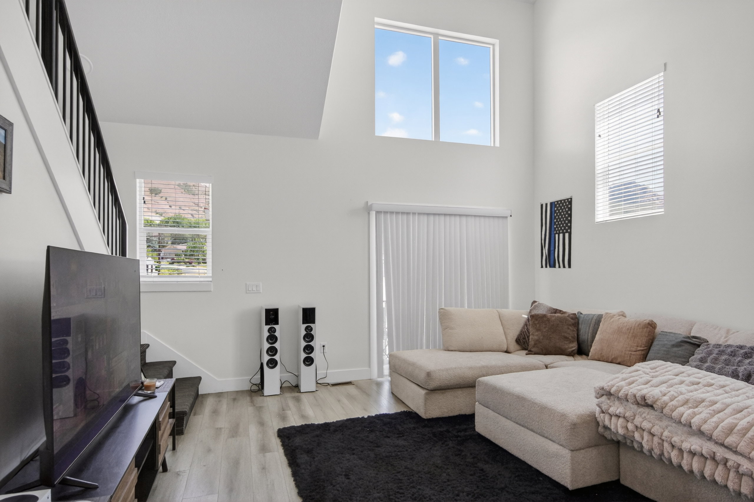 Spacious living area featuring high ceilings, white walls, wood-finish flooring, and a sliding glass door with vertical blinds