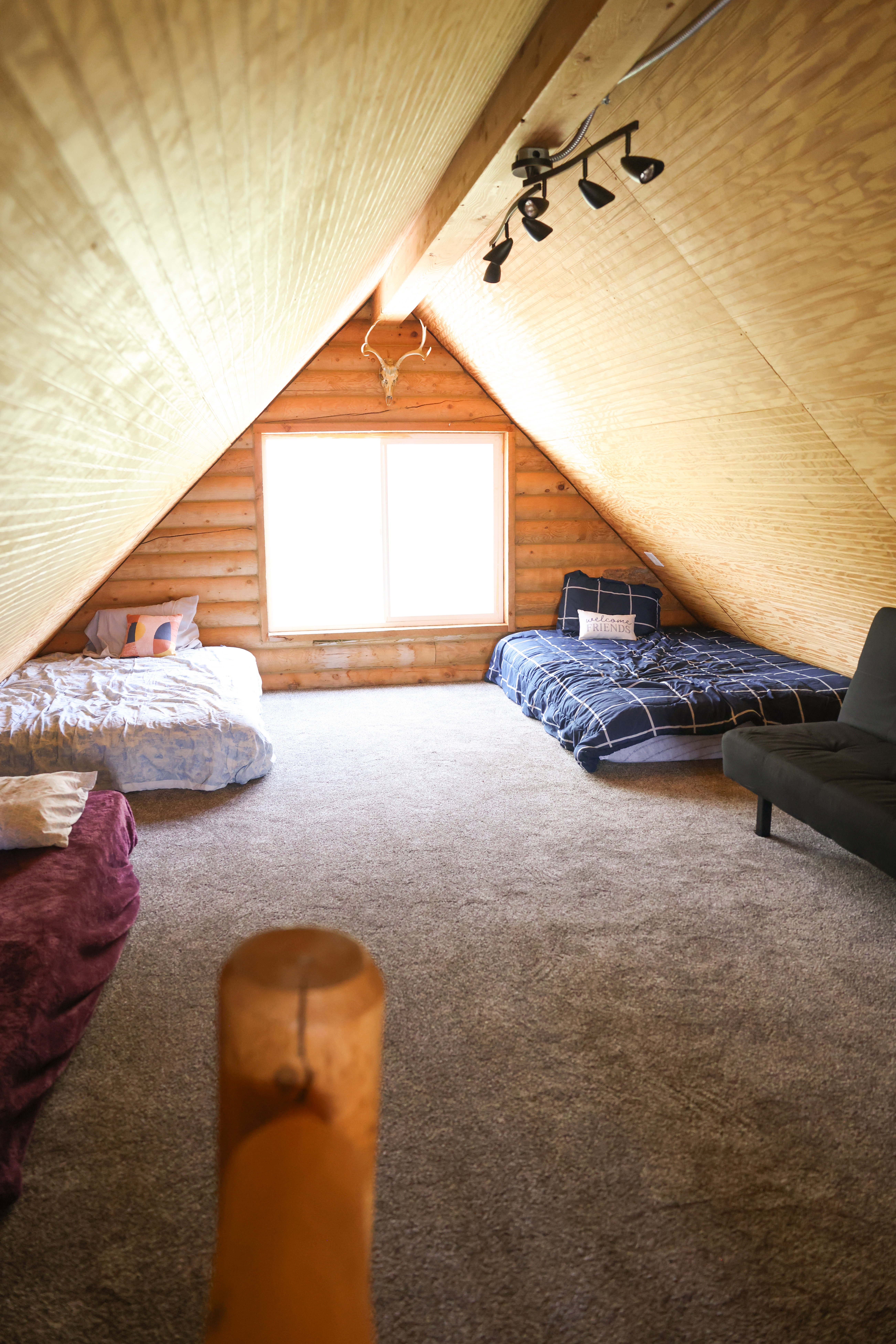 Spacious attic room featuring sloped wood-paneled ceilings, a bright central window, track lighting, and soft carpet flooring