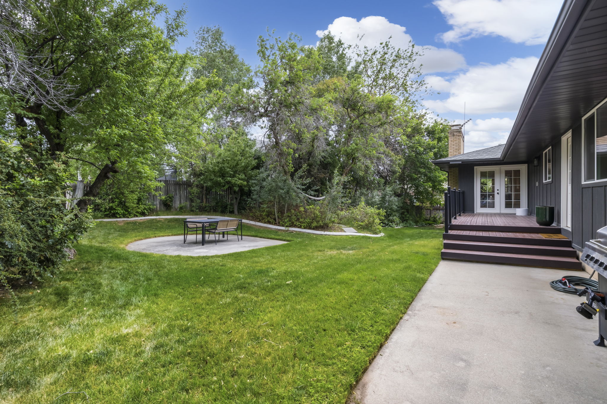 Expansive backyard featuring a manicured lawn, a circular concrete patio, mature trees, a multi-level wood deck, and French doors leading into the property