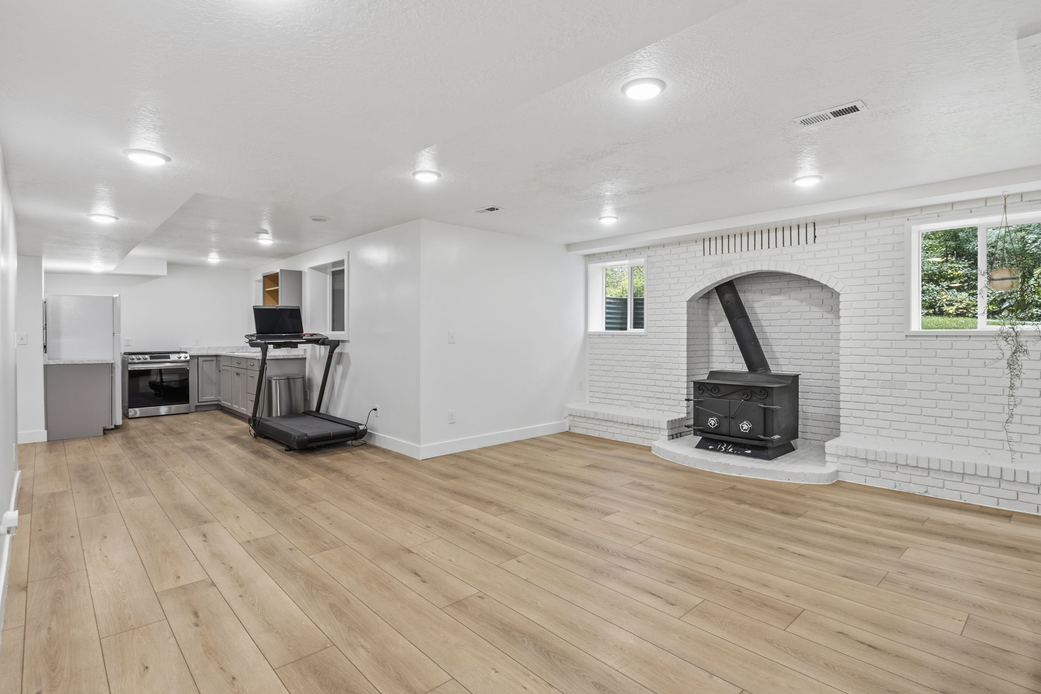 Spacious open-concept living area featuring wood-finish flooring, recessed lighting, and a white brick-faced hearth with a freestanding stove