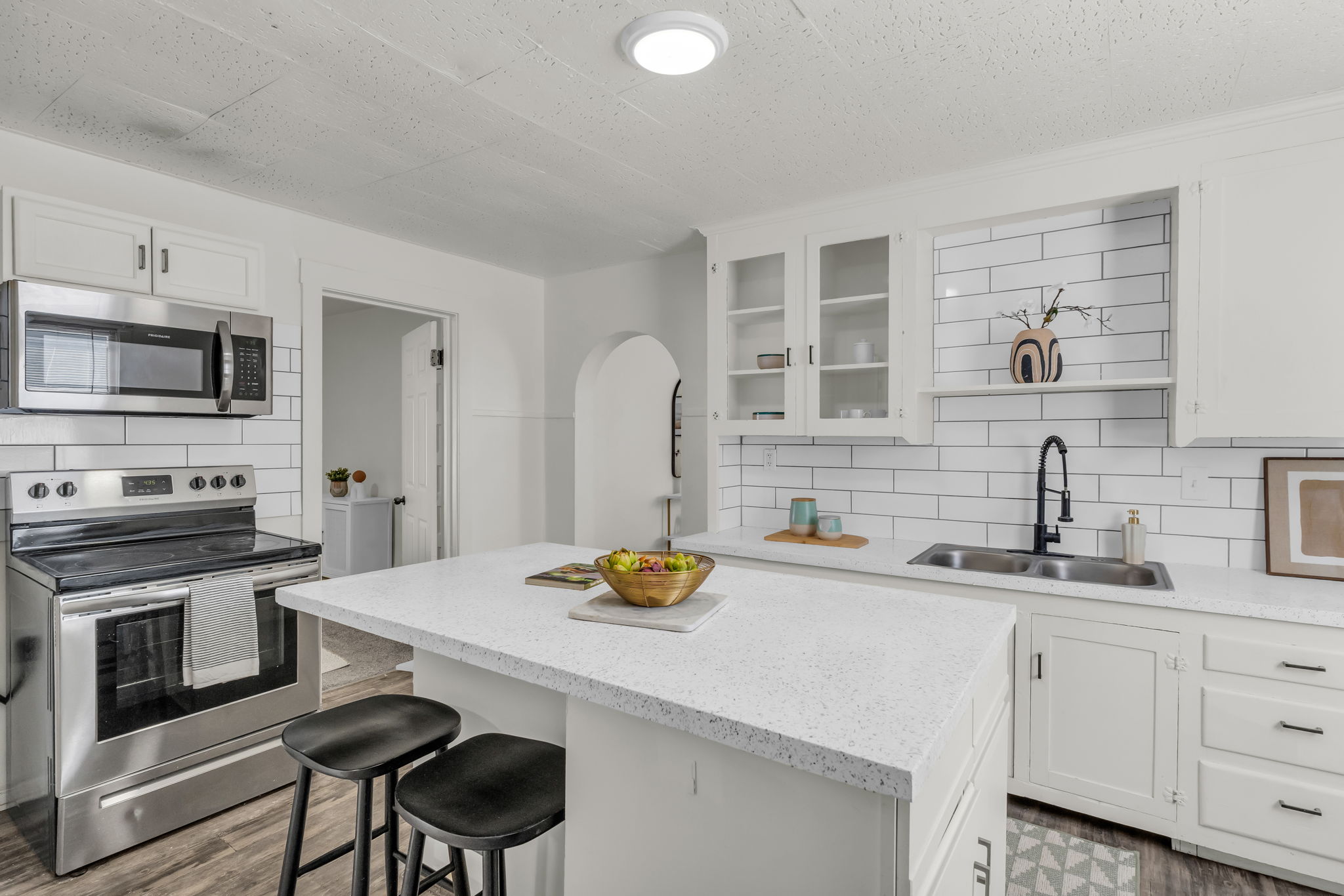 Modern kitchen featuring a central island with a light-colored countertop, stainless steel appliances, a subway tile backsplash, white cabinetry with glass-front uppers, and wood-finish flooring