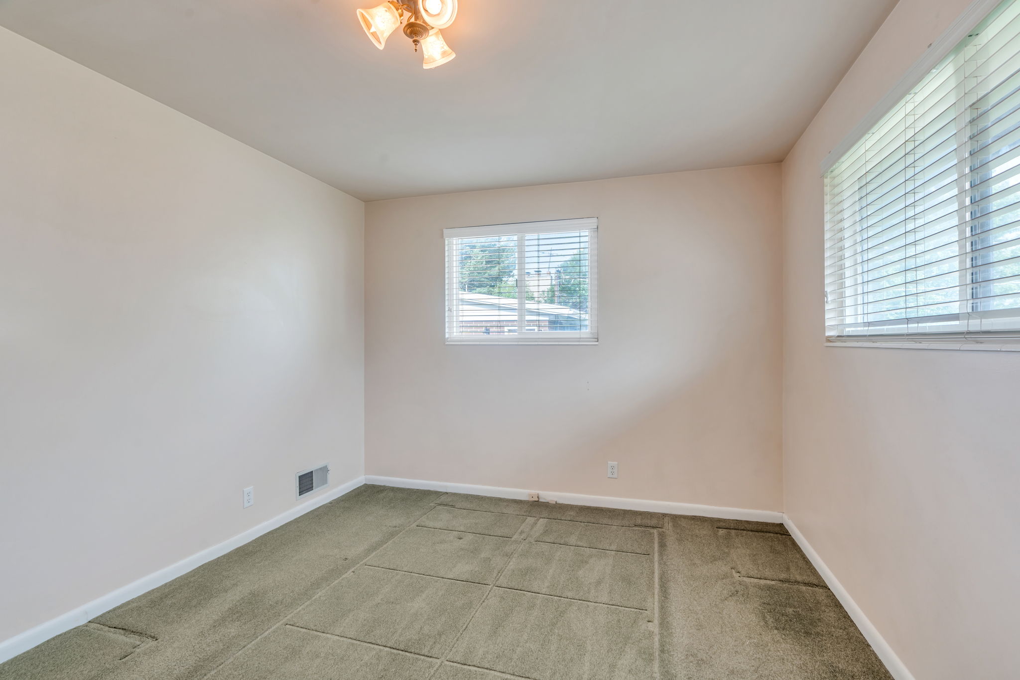 Room featuring two windows with blinds, light beige walls, and green carpet
