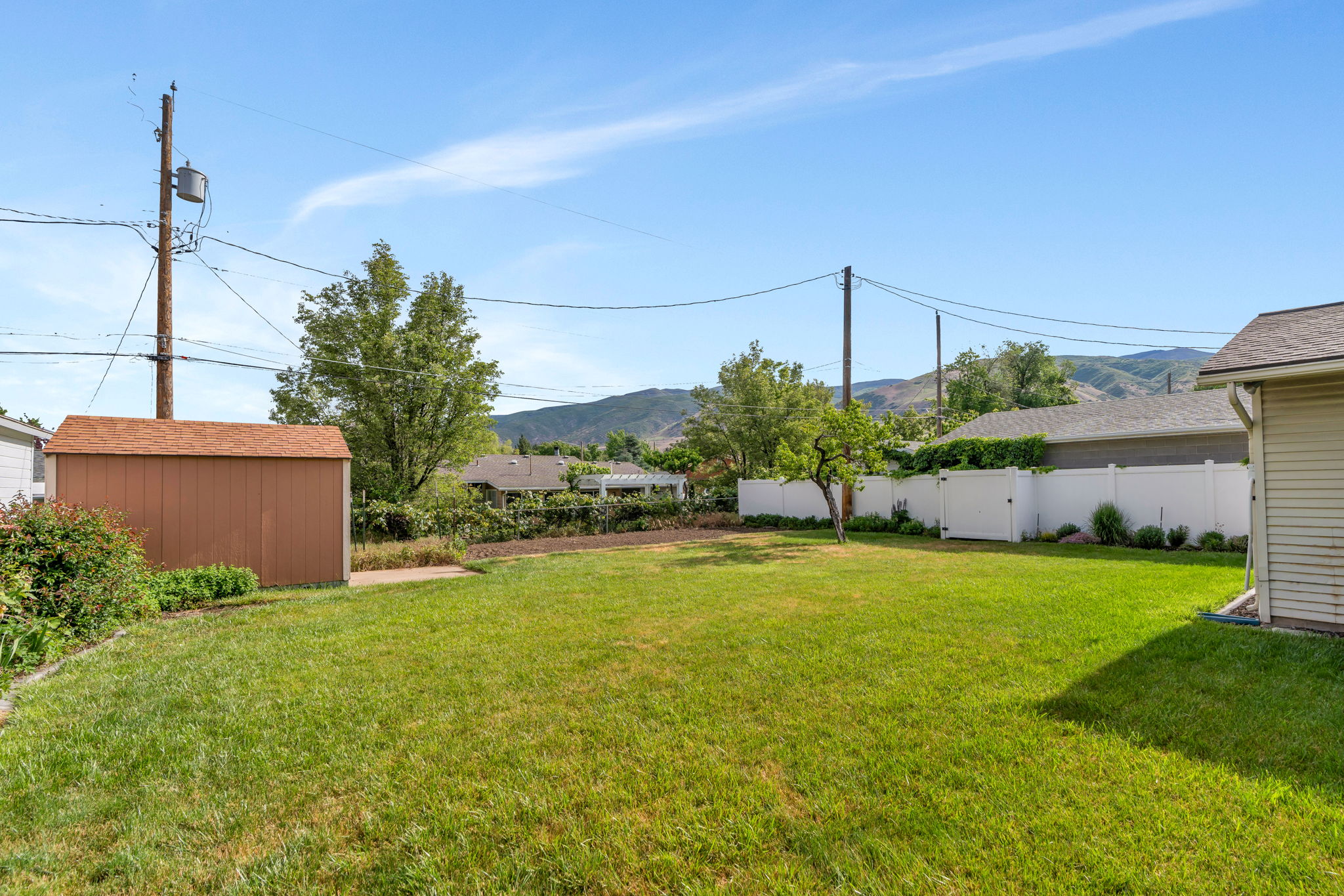 Expansive backyard featuring a manicured lawn, a brown storage shed with a shingle roof, a white privacy fence, and mature trees