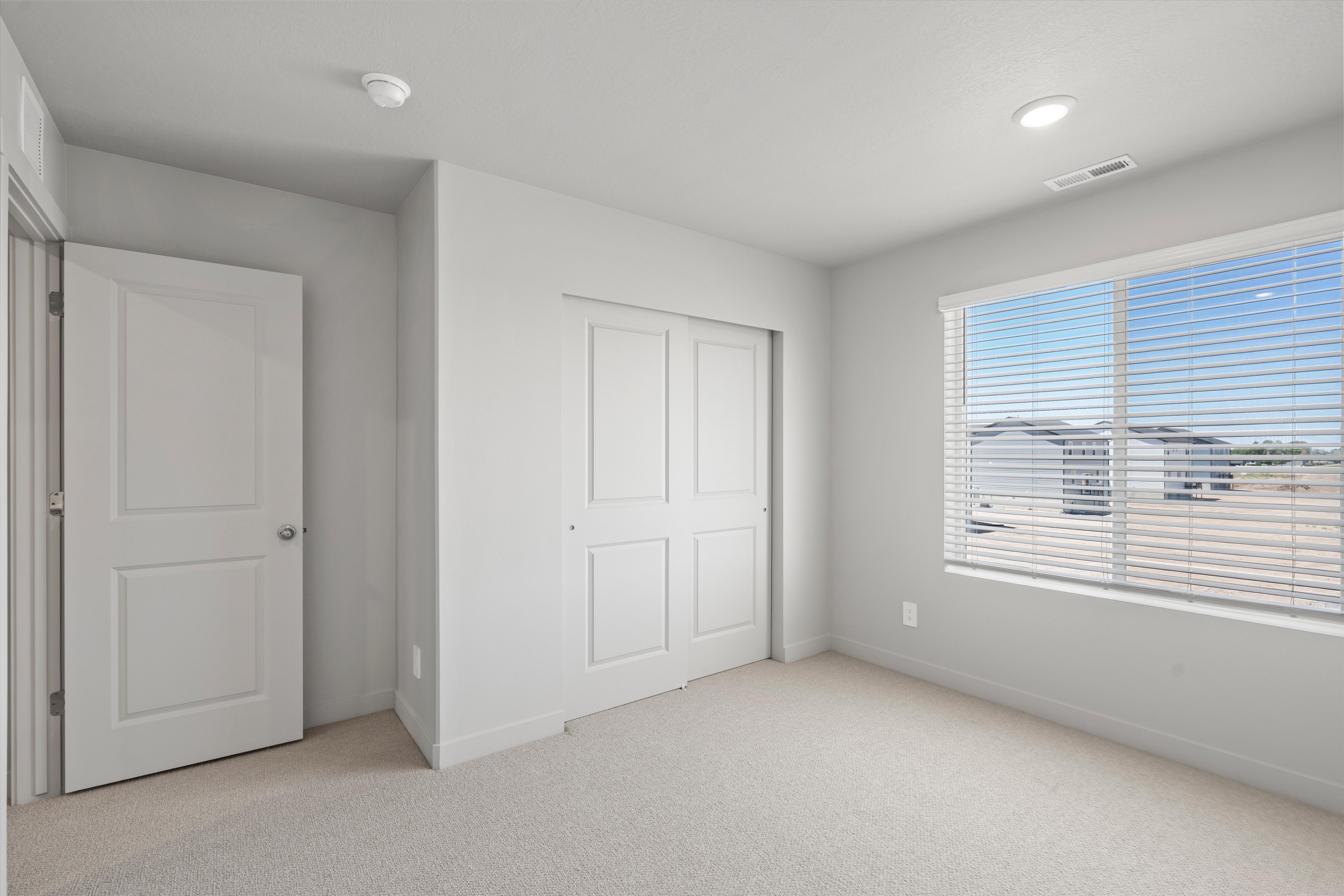 Bright room with light gray walls, plush carpeting, a large window with horizontal blinds, a sliding two-panel closet door, and an overhead recessed light fixture