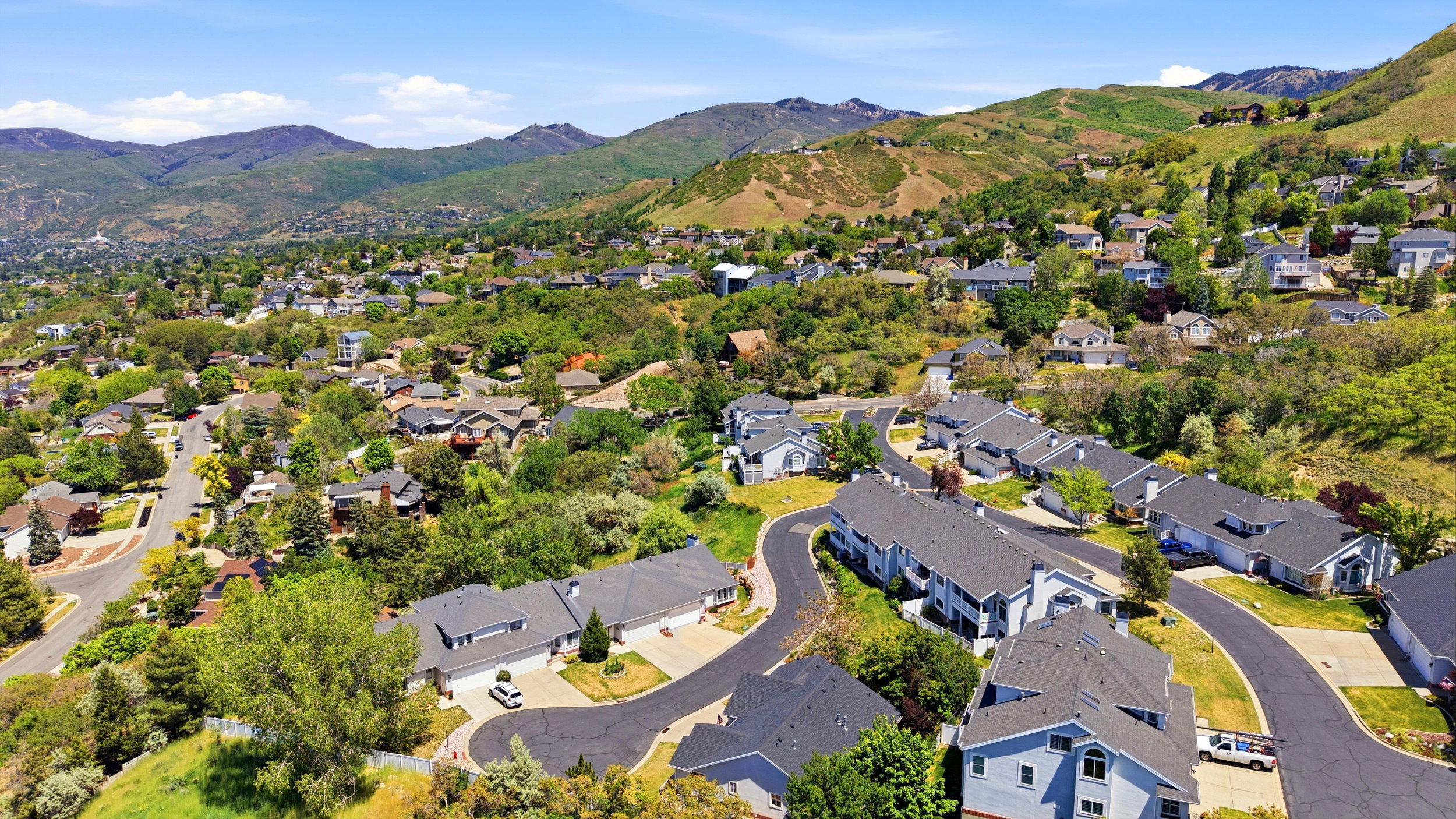Residential community featuring a variety of architectural styles, mature trees, paved streets, and a mountainous backdrop under a clear sky