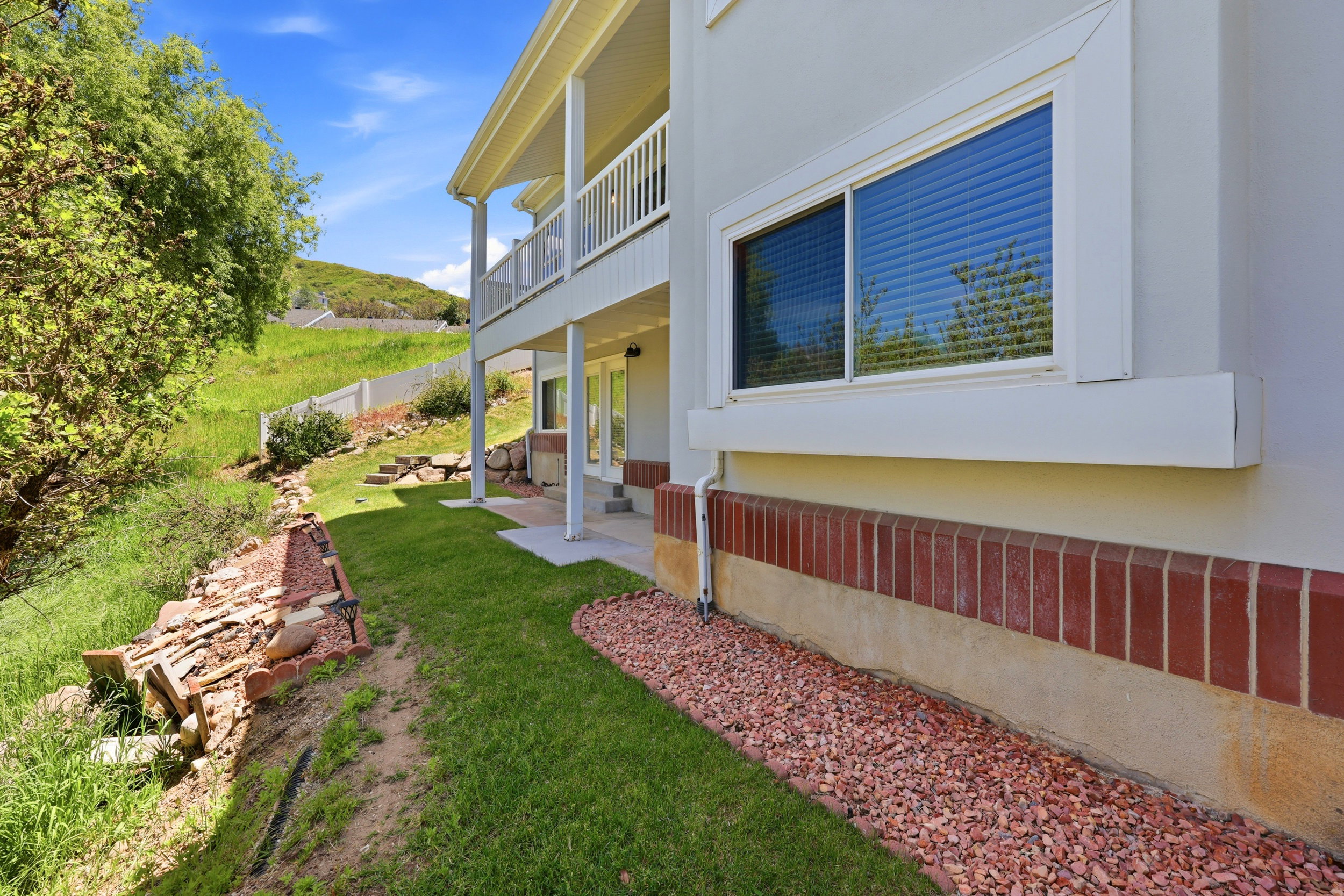 Exterior featuring a lower-level patio with overhead balcony, red brick foundation, and light stucco siding
