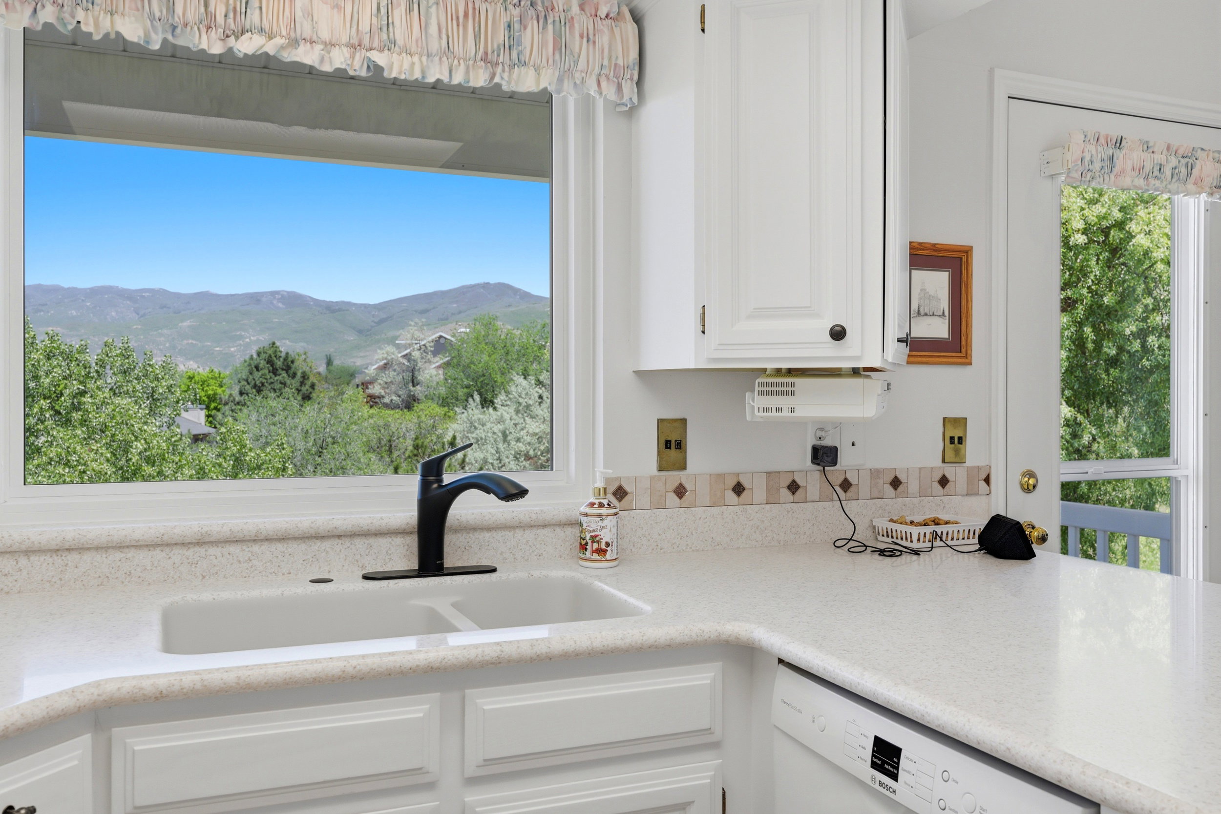 Kitchen window with a mountain backdrop, featuring a double basin sink, black faucet, light-colored countertops, and white cabinetry