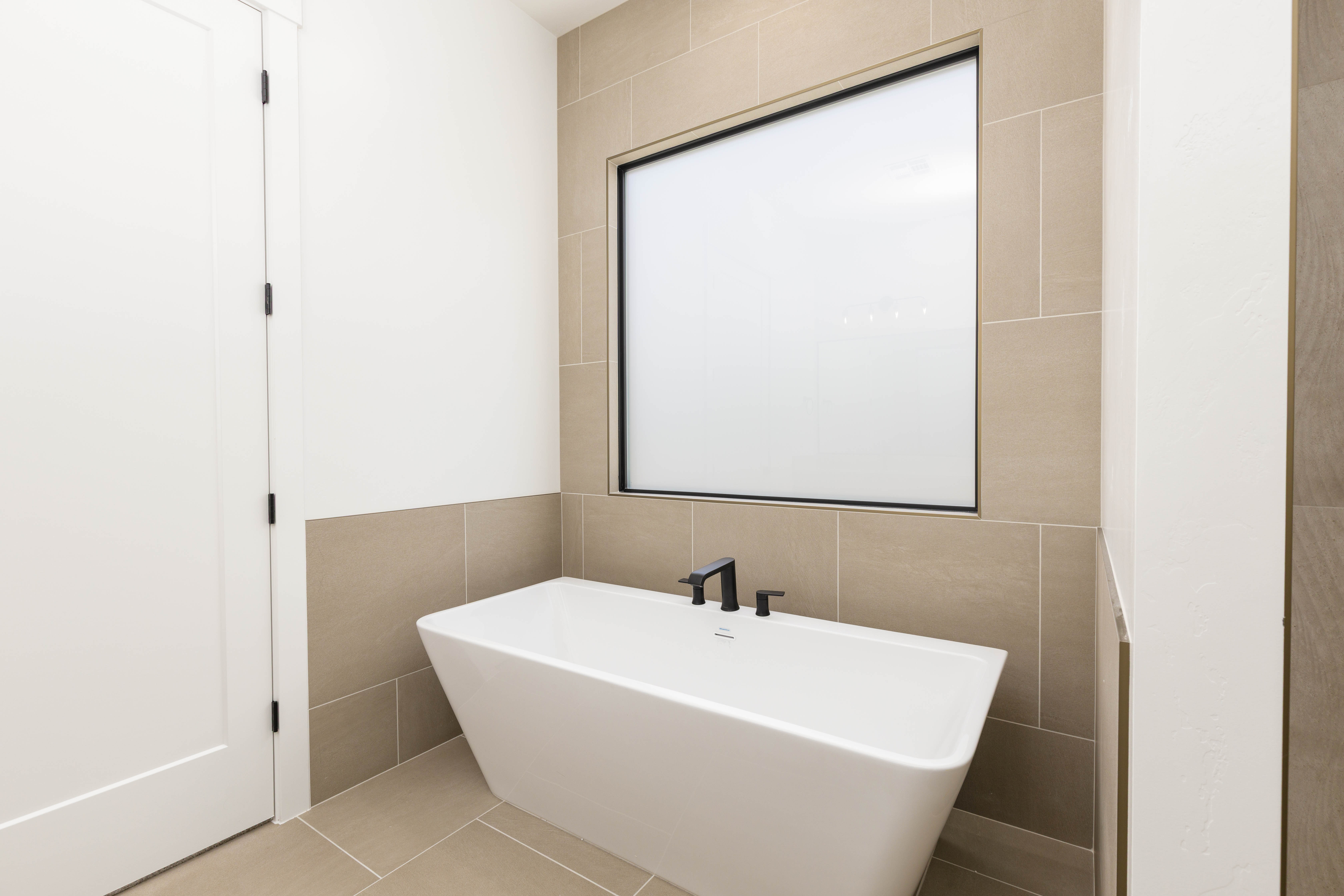 Freestanding soaking tub with matte black fixtures, set against a tiled wall with an integrated frosted window