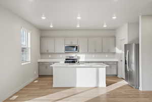 Modern kitchen featuring a central island, light wood-finish flooring, recessed lighting, stainless steel appliances, and light gray cabinetry with white countertops