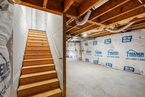 Unfinished basement space featuring exposed wooden ceiling joists, concrete flooring, and an open staircase with wooden treads
