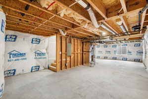 Unfinished basement space featuring exposed wooden joists, concrete flooring, foundational walls with insulation, and visible utility conduits