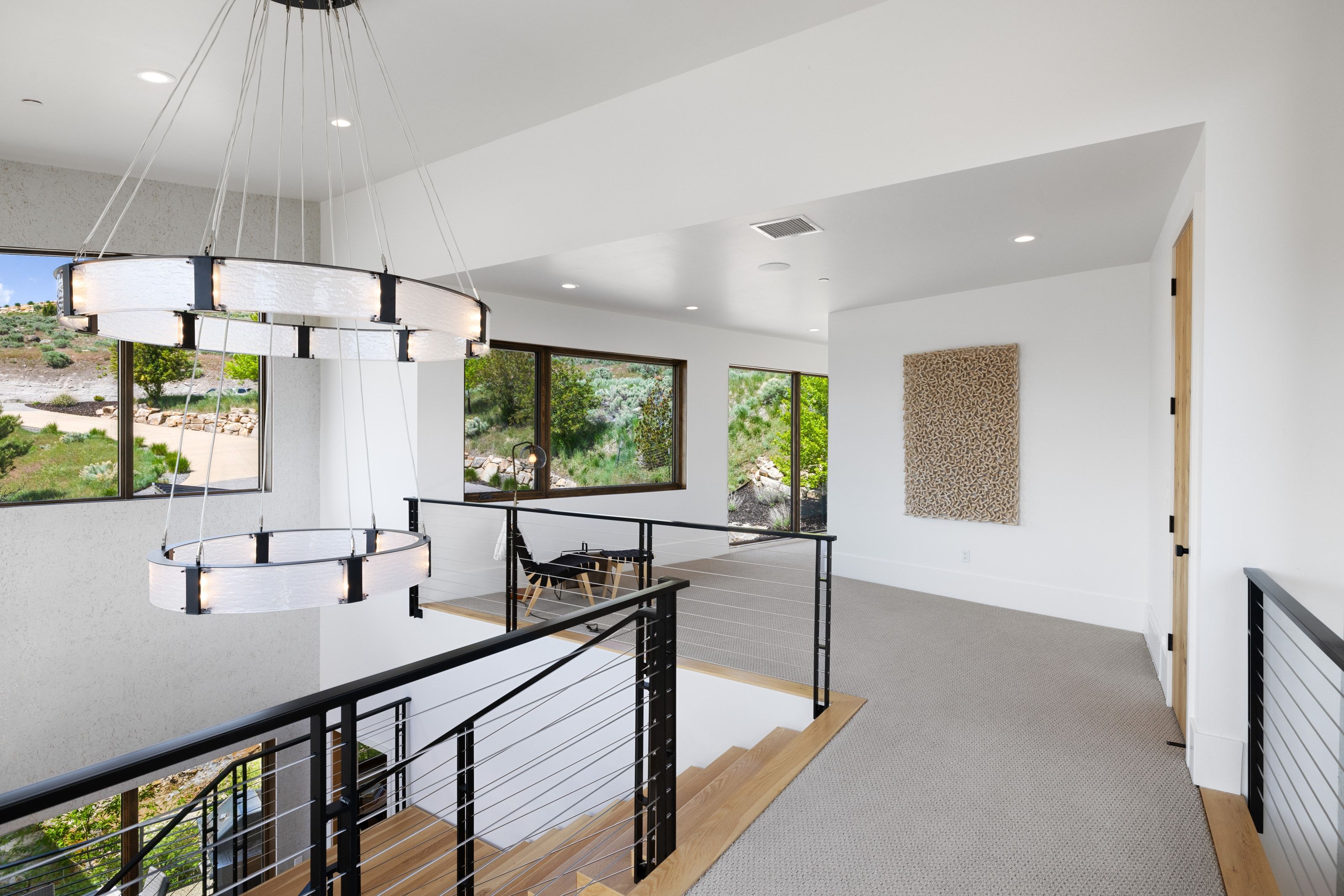 Contemporary two-tiered chandelier illuminating a landing with light wood-finish stairs and cable railing
