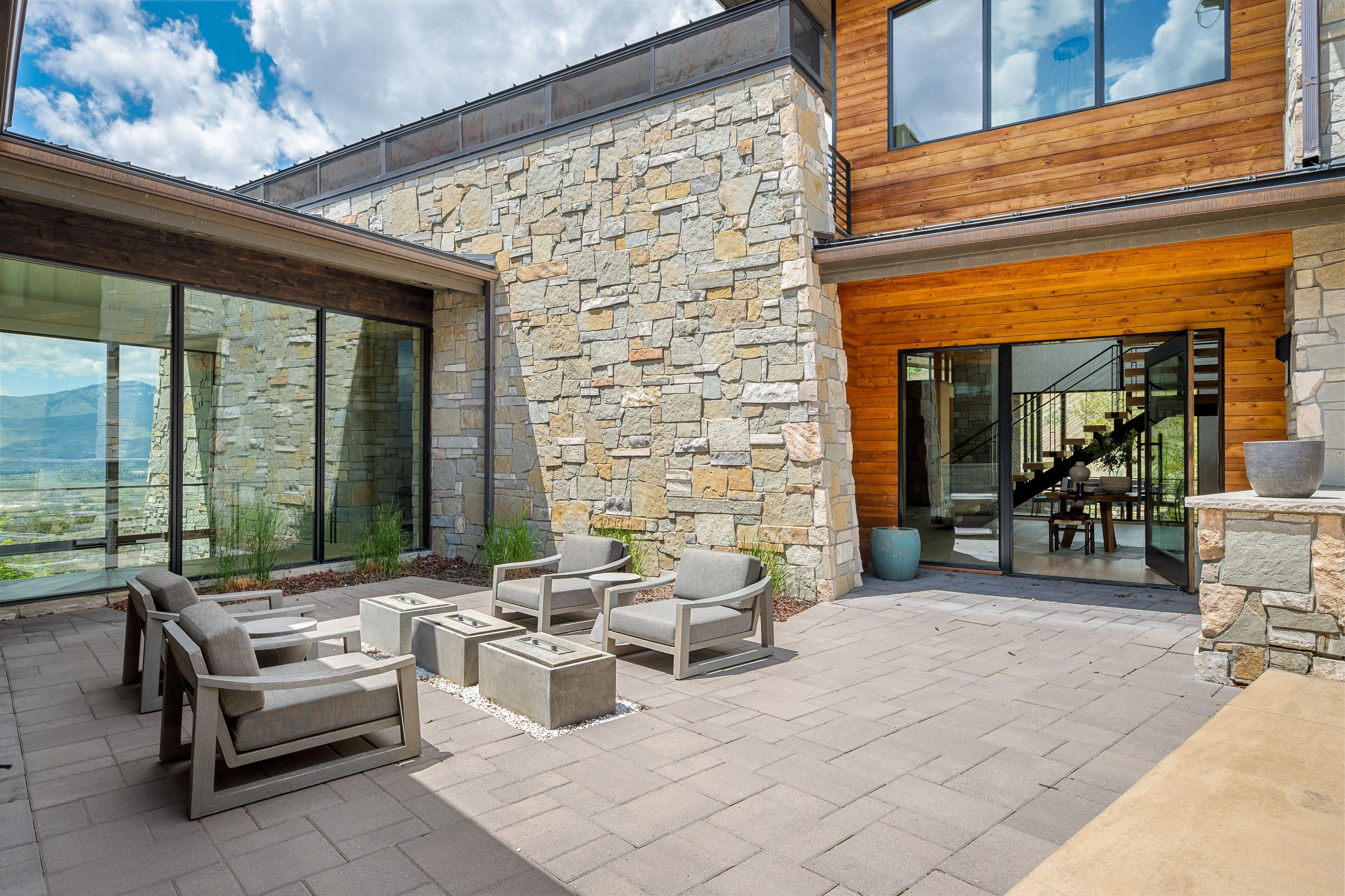 Expansive outdoor patio with large format pavers, stone veneer exterior, wood paneling accents, floor-to-ceiling windows, and multi-panel sliding glass doors