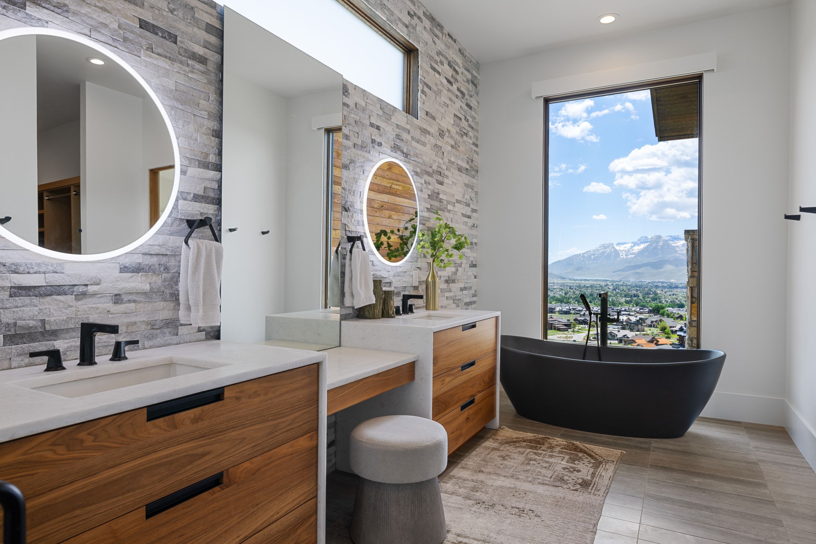 Modern bathroom featuring a black freestanding bathtub with floor-mounted faucet, a vanity with wood-finish cabinetry, integrated sinks, and backlit circular mirrors, and a stone accent wall