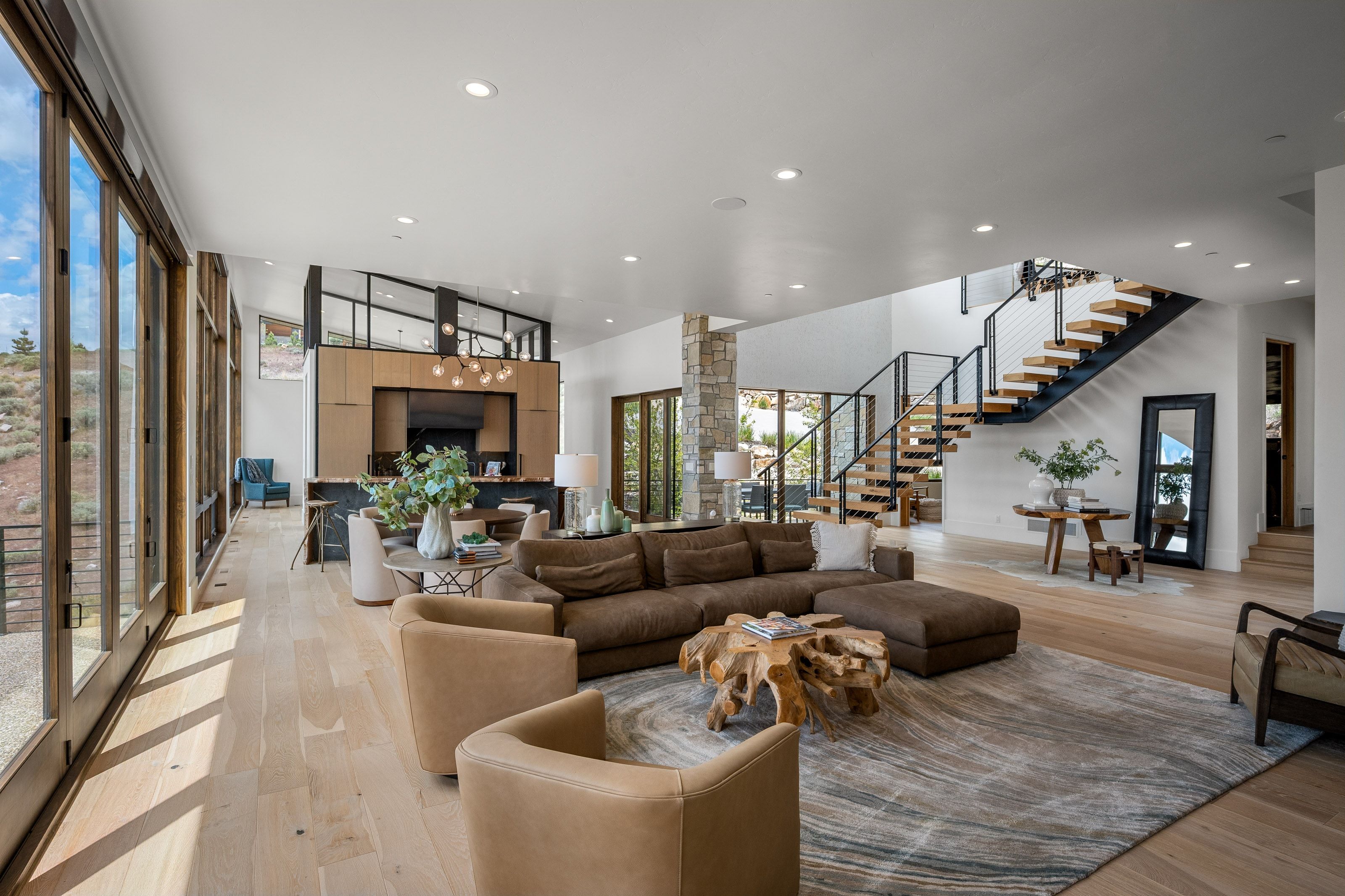 Spacious great room featuring light wood-finish flooring, a contemporary floating staircase with metal railings, a stone-clad pillar, recessed ceiling lighting, and expansive glass doors