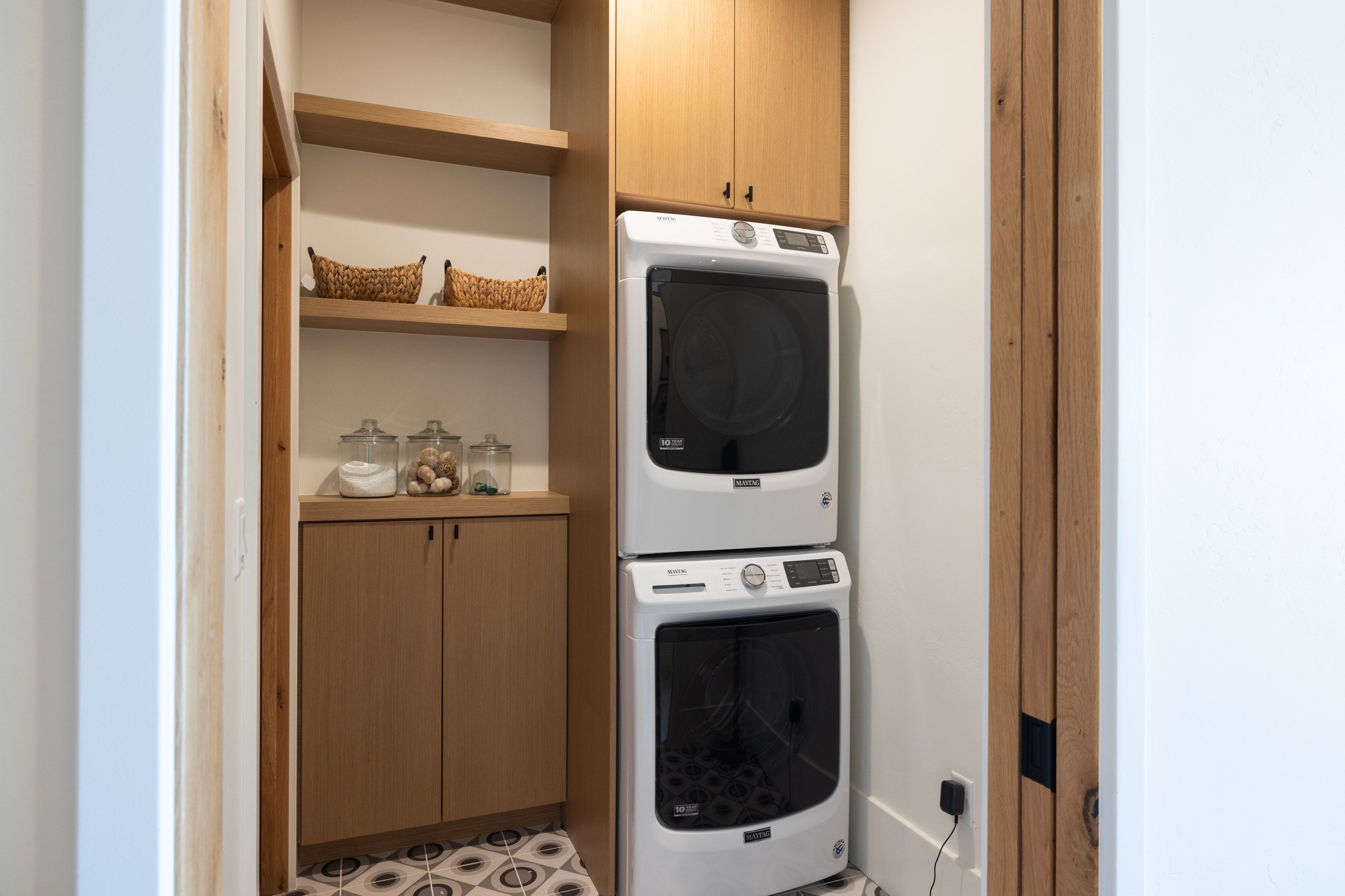 Dedicated laundry area featuring a stacked washer and dryer, wood-finish cabinetry, open shelving, and patterned floor tiling