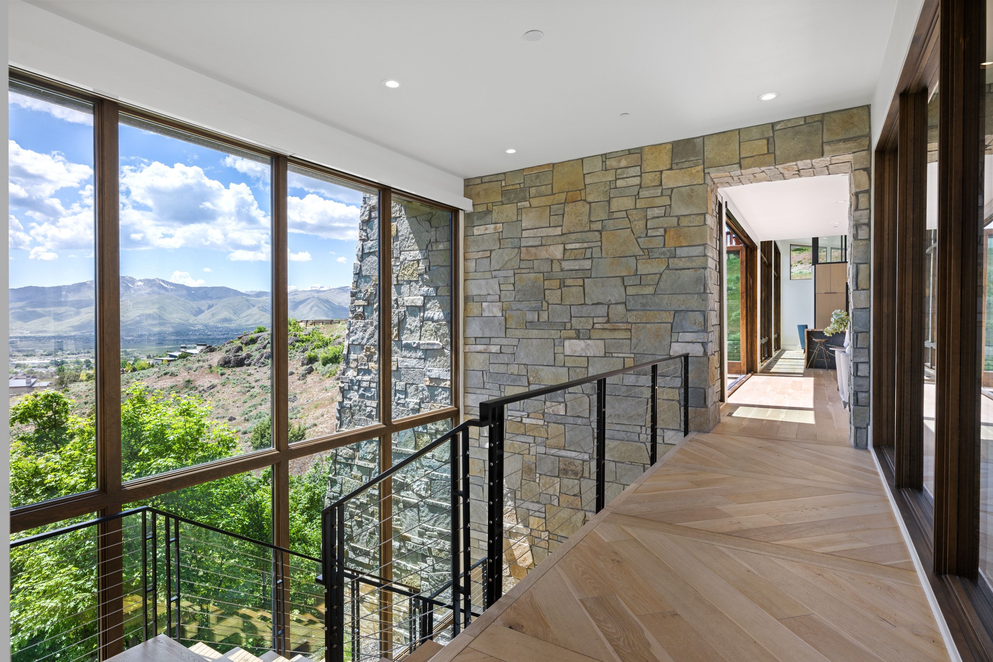 Bright hallway with expansive floor-to-ceiling windows, stone accent wall, chevron wood-finish flooring, recessed lighting, and a contemporary metal and cable railing system