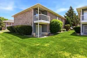 Exterior featuring brick siding, a private balcony with white railings, a ground-level patio, mature shrubbery, and a manicured lawn